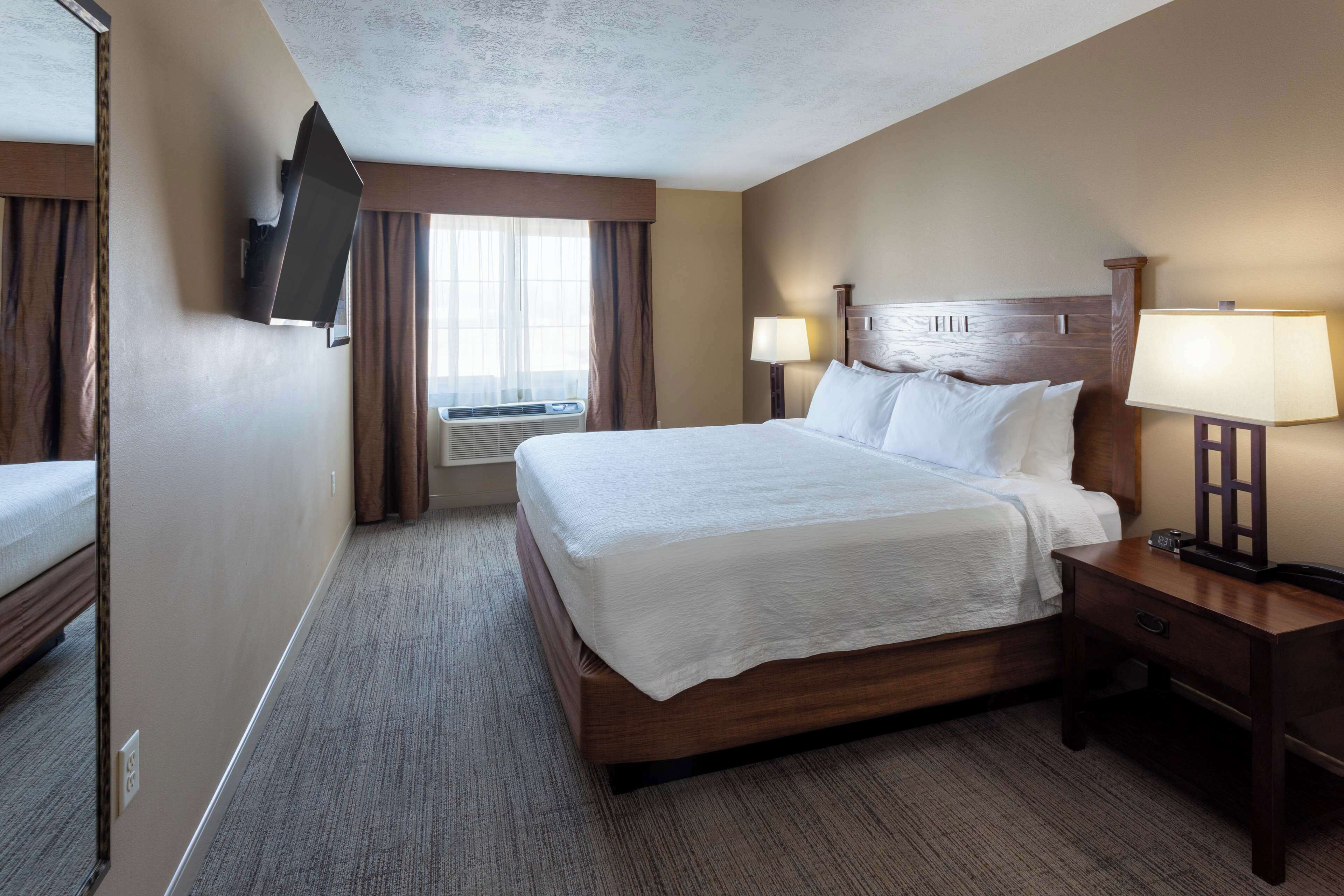 A hotel room with a large bed, a flat-screen TV mounted on the wall, a bedside table with a lamp, and a window with curtains.