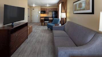 A hotel room with a large flat-screen TV, a sofa, a chair, a small table, a lamp, a framed picture on the wall, and a kitchenette area.