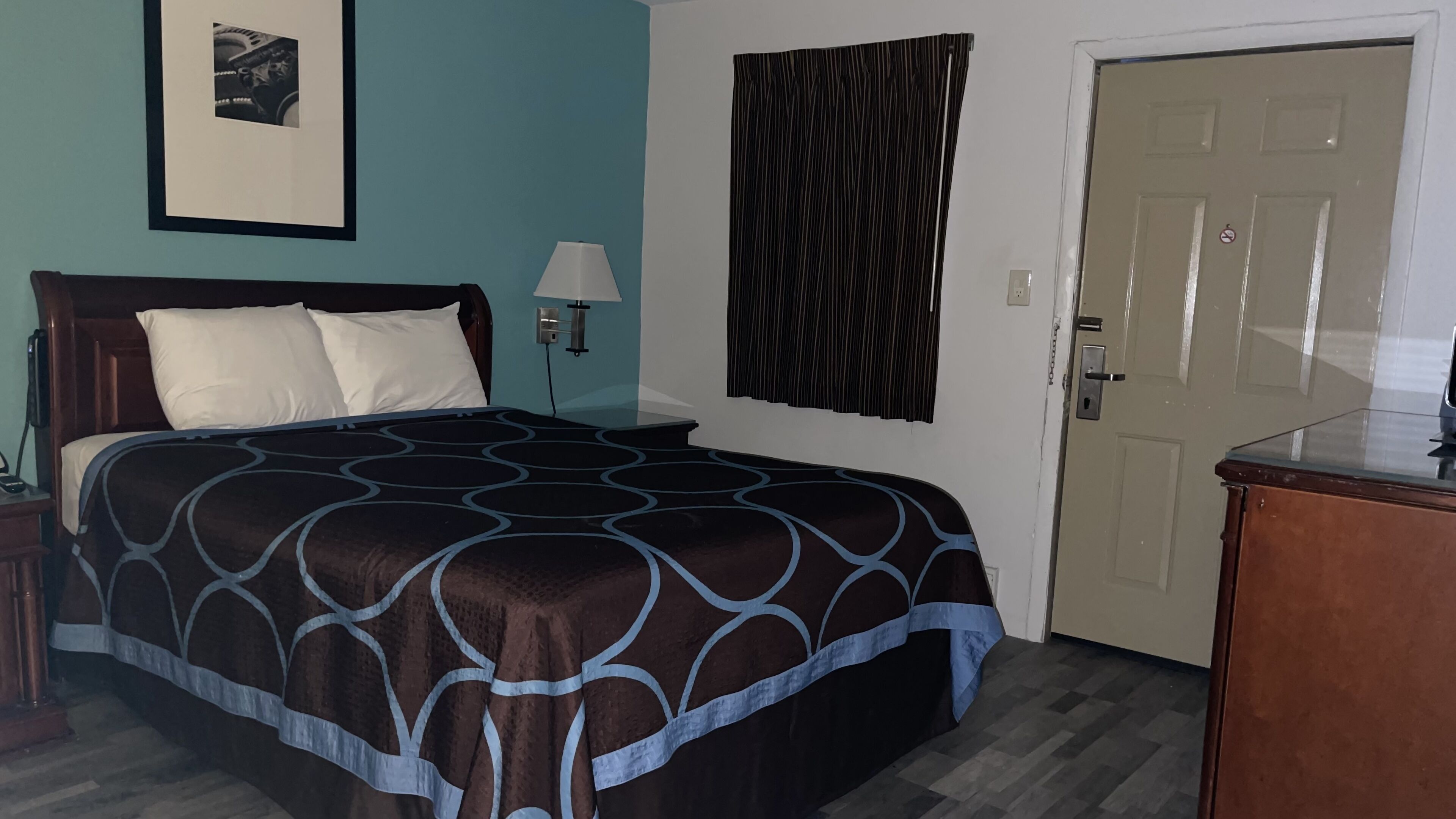 A bedroom with a bed, a nightstand, a lamp, a door, and a framed picture on the wall.