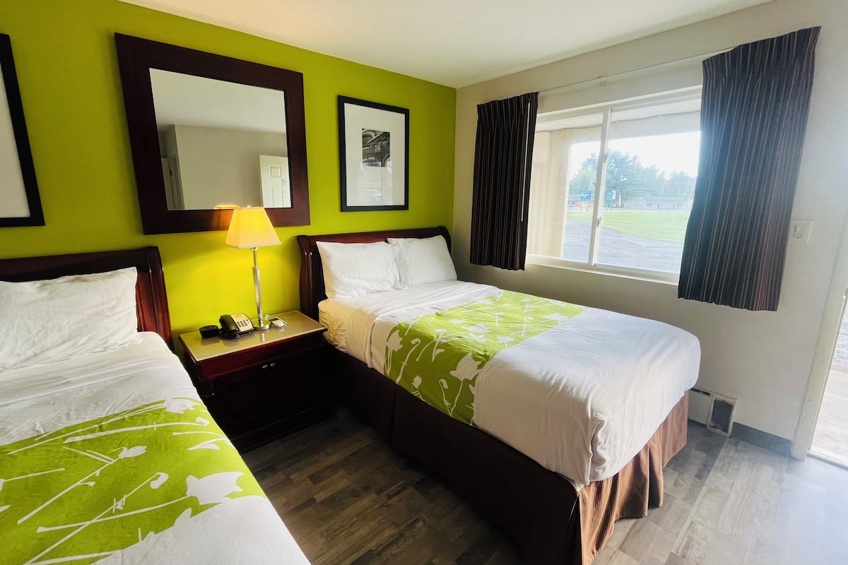 A hotel room with two beds, a bedside table with a lamp, a window with curtains, and a mirror on the wall.
