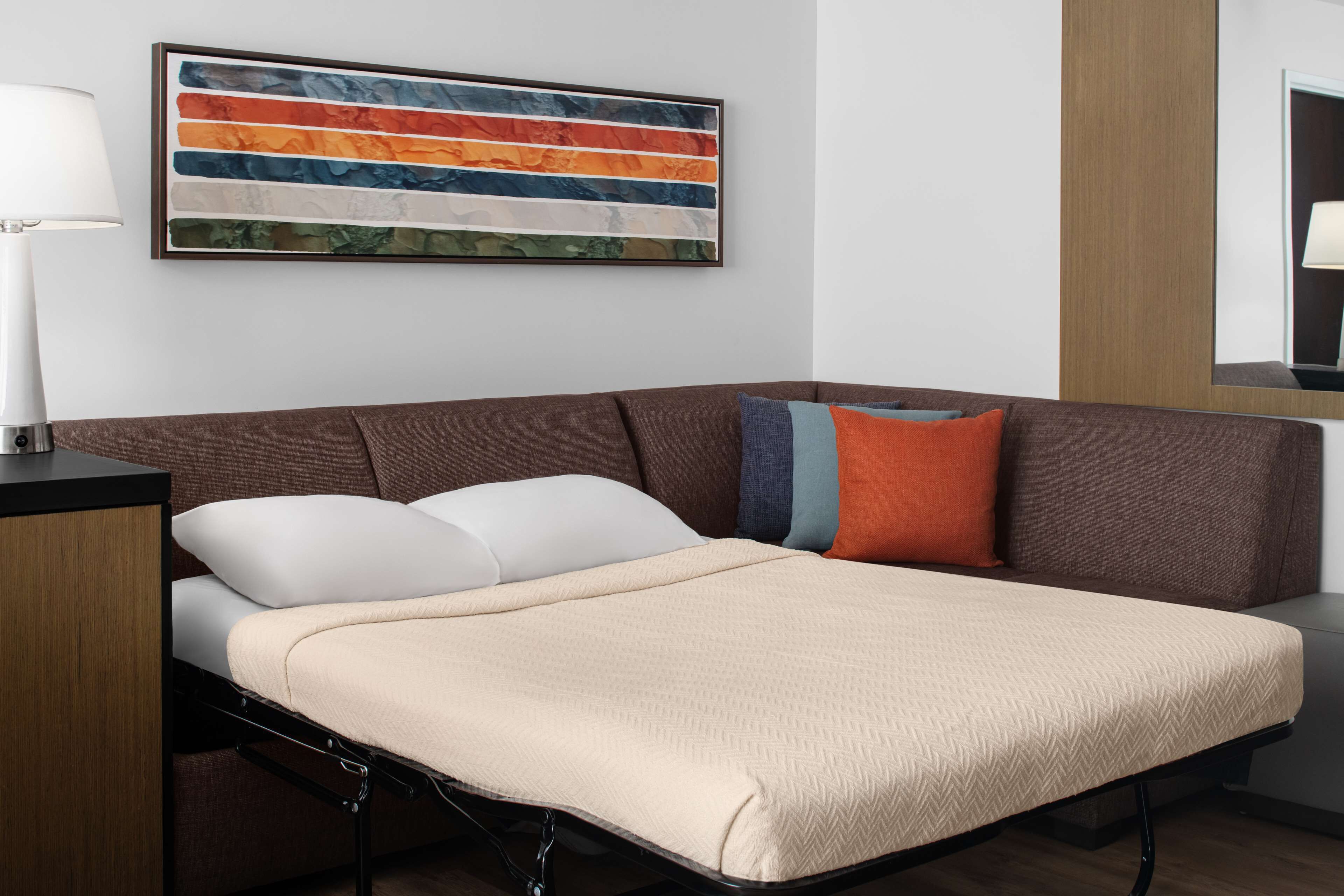 A modern room with a sofa bed, a side table, a lamp, and a colorful abstract painting.