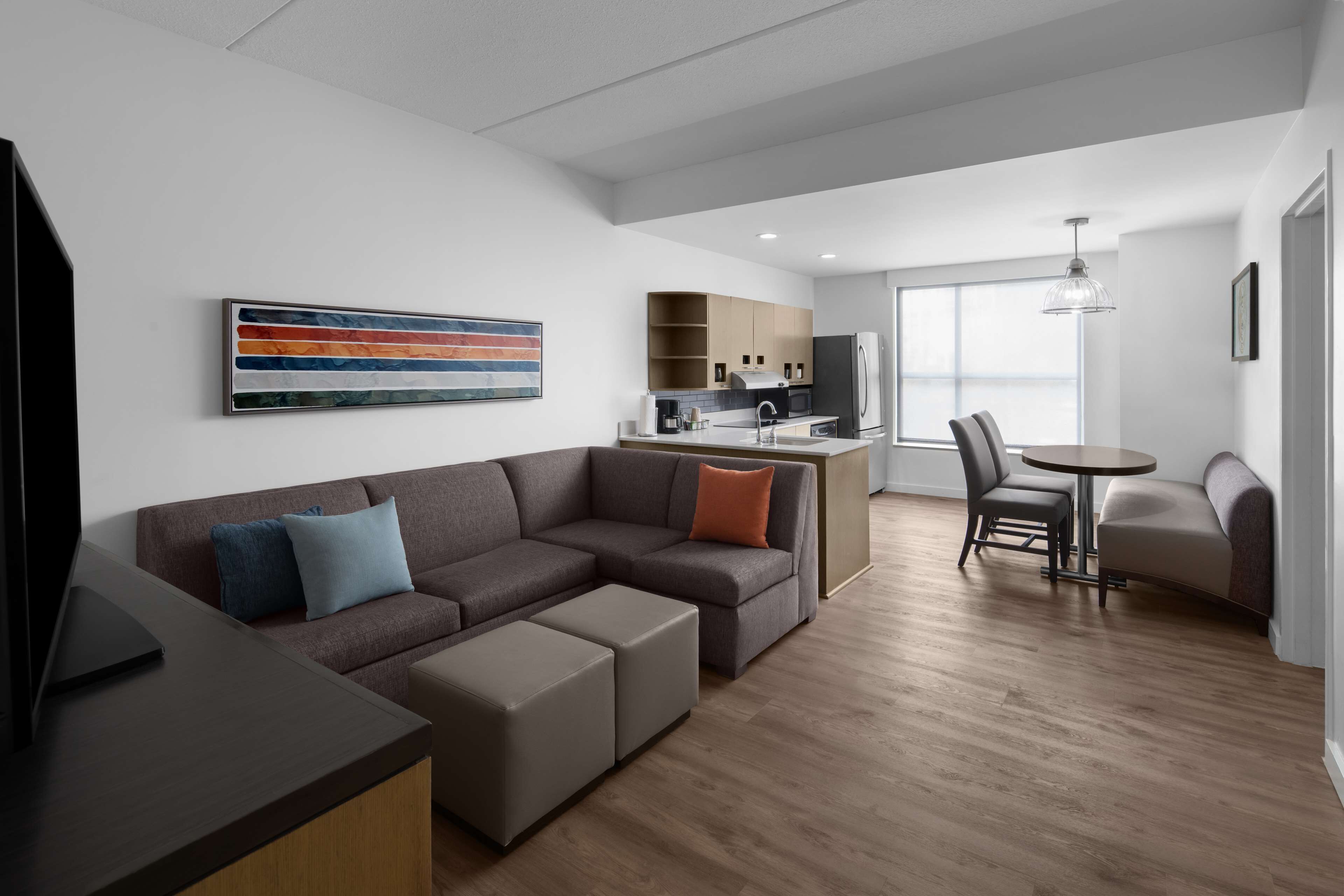 A modern living room with a sectional sofa, ottomans, a dining table with chairs, a kitchen area, and a large window.
