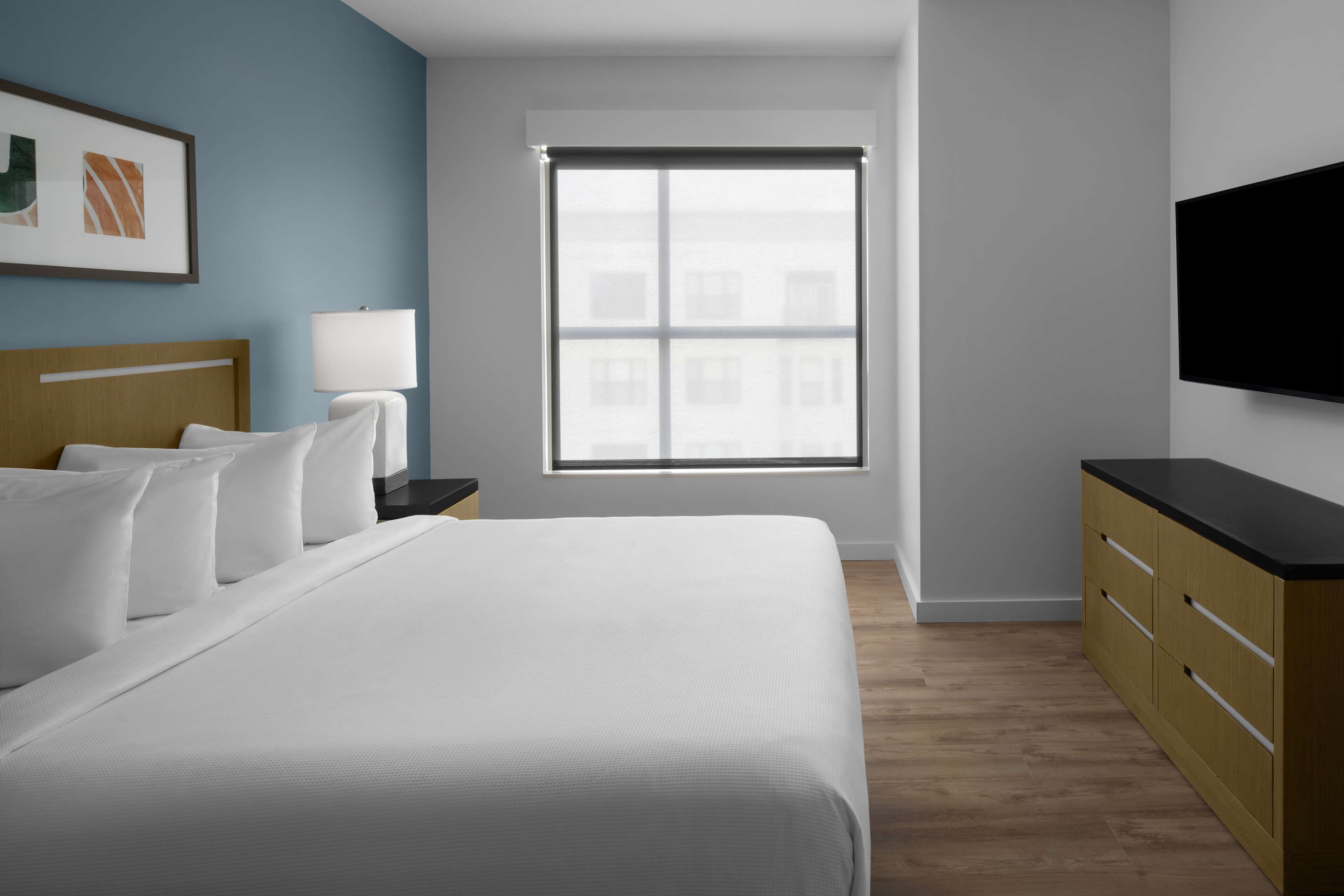 Une chambre d’hôtel avec un grand lit, une table de chevet avec une lampe, une commode, un téléviseur fixé au mur et une fenêtre avec des stores.