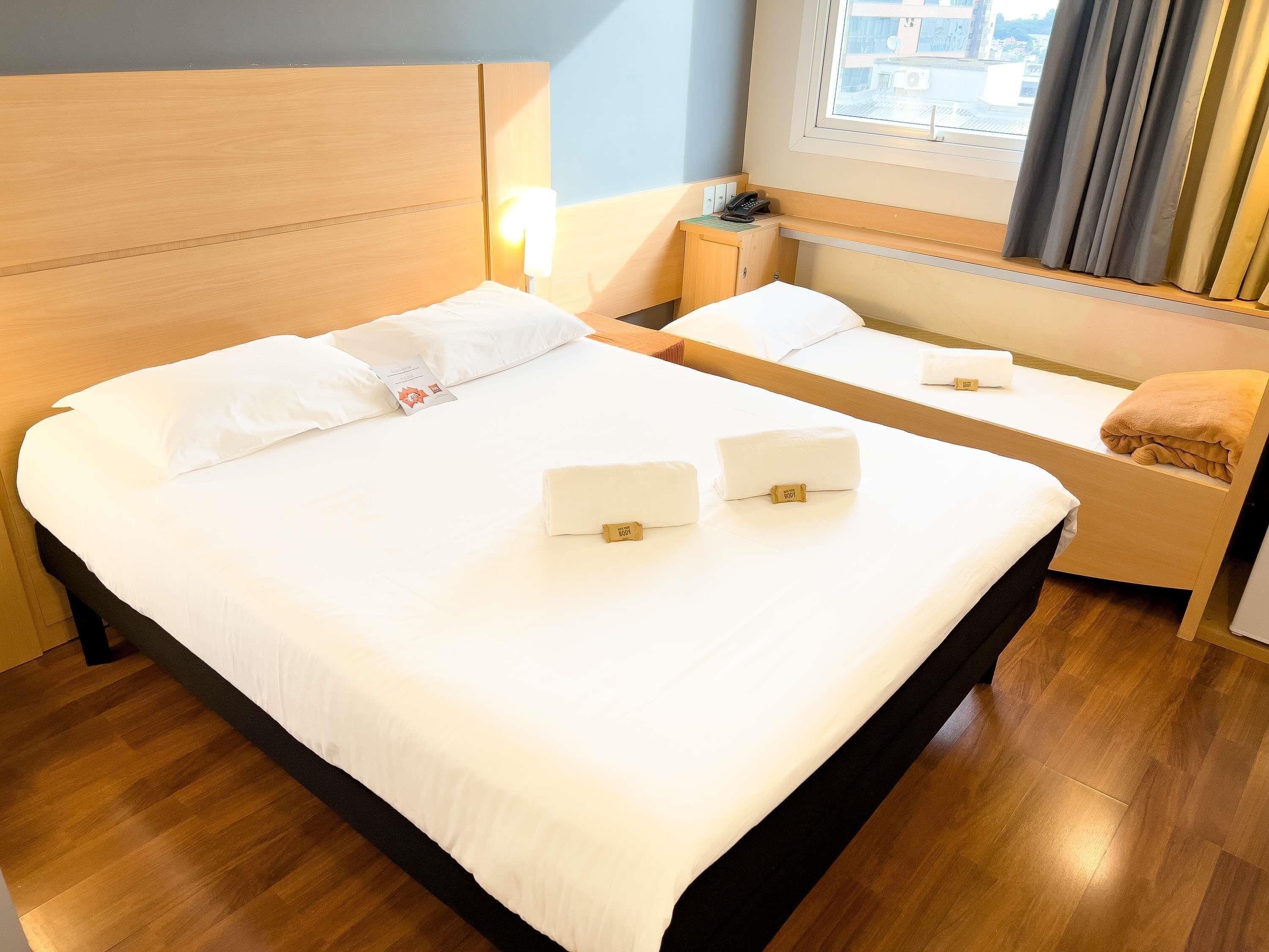 A hotel room with two beds, a bedside table with a lamp, and a window with curtains.