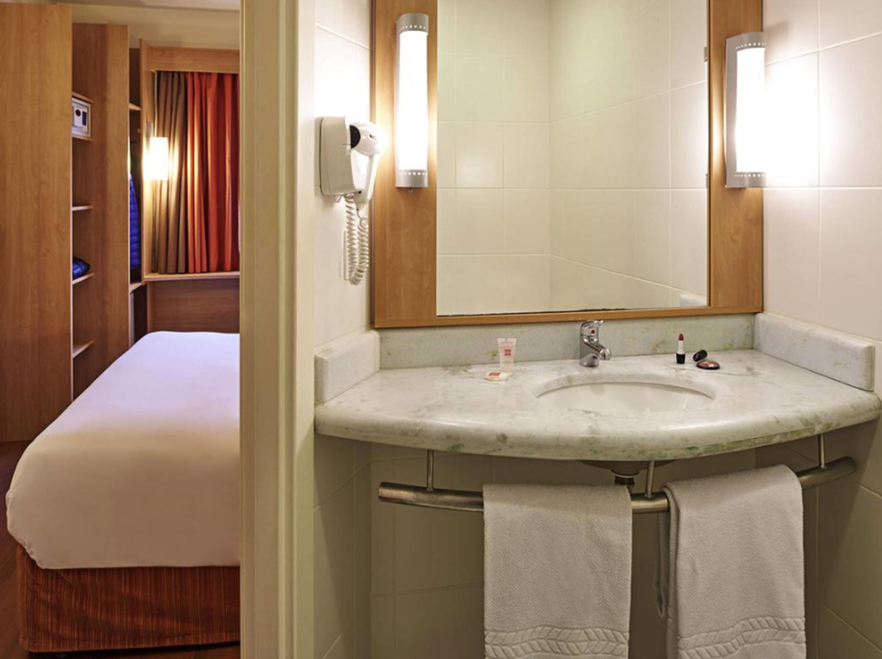 A hotel bathroom with a large mirror, a sink, and a toilet. There is a bed visible in the background.