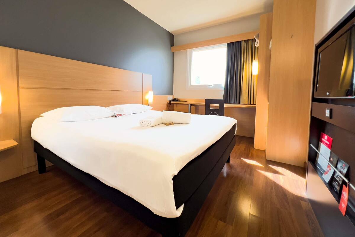 A hotel room with a large bed, a desk, a chair, a TV, and a window with curtains.