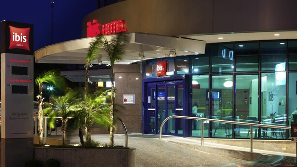 A hotel entrance with a sign for ibis hotels, a glass door entrance, and a palm tree in the foreground. - ibis Passo Fundo Centro Hotel (Passo Fundo)