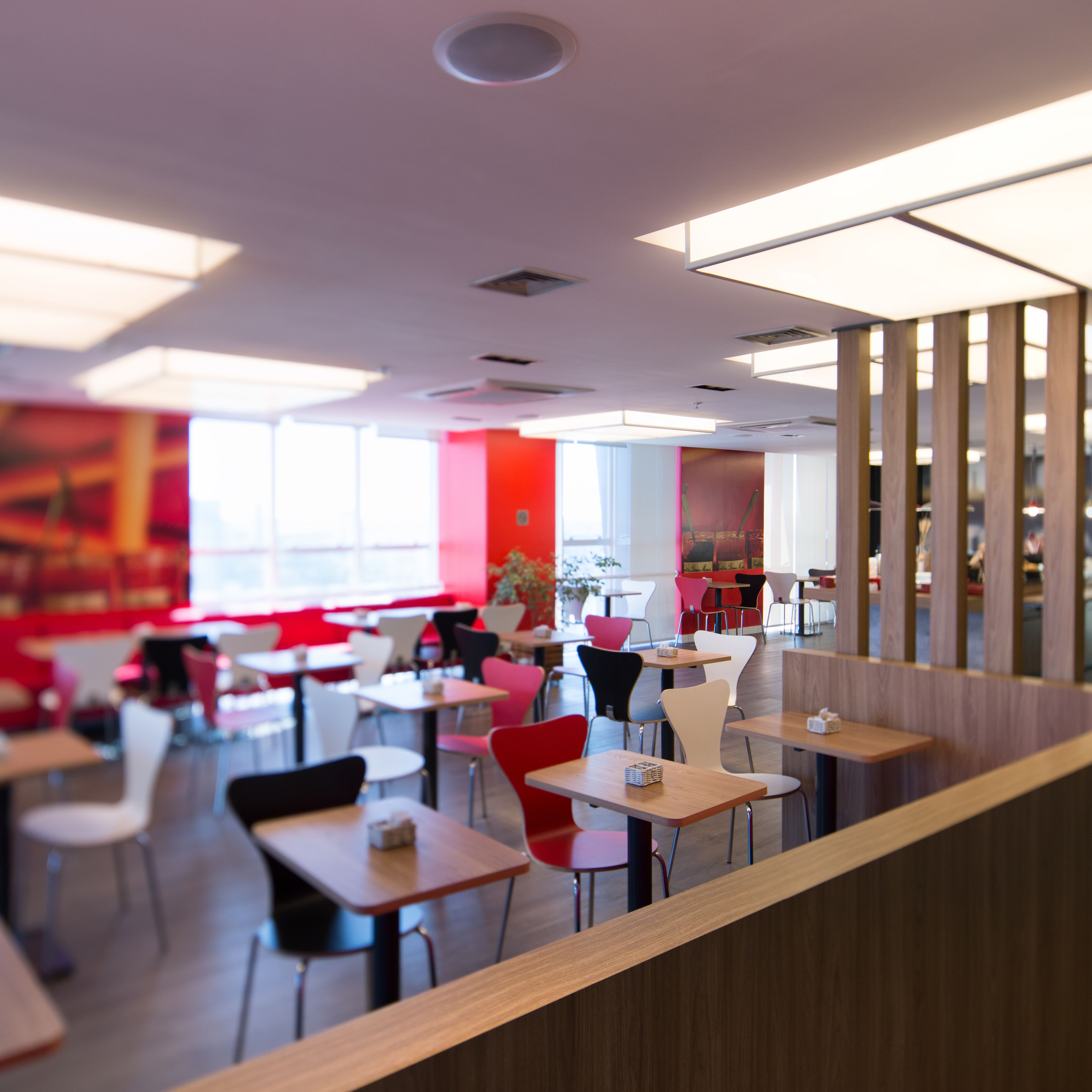 Interior modern d'un restaurant amb diverses taules i caires, un sostre alt amb il·luminació empotrada i una paret d'accent vermella i vibrant.