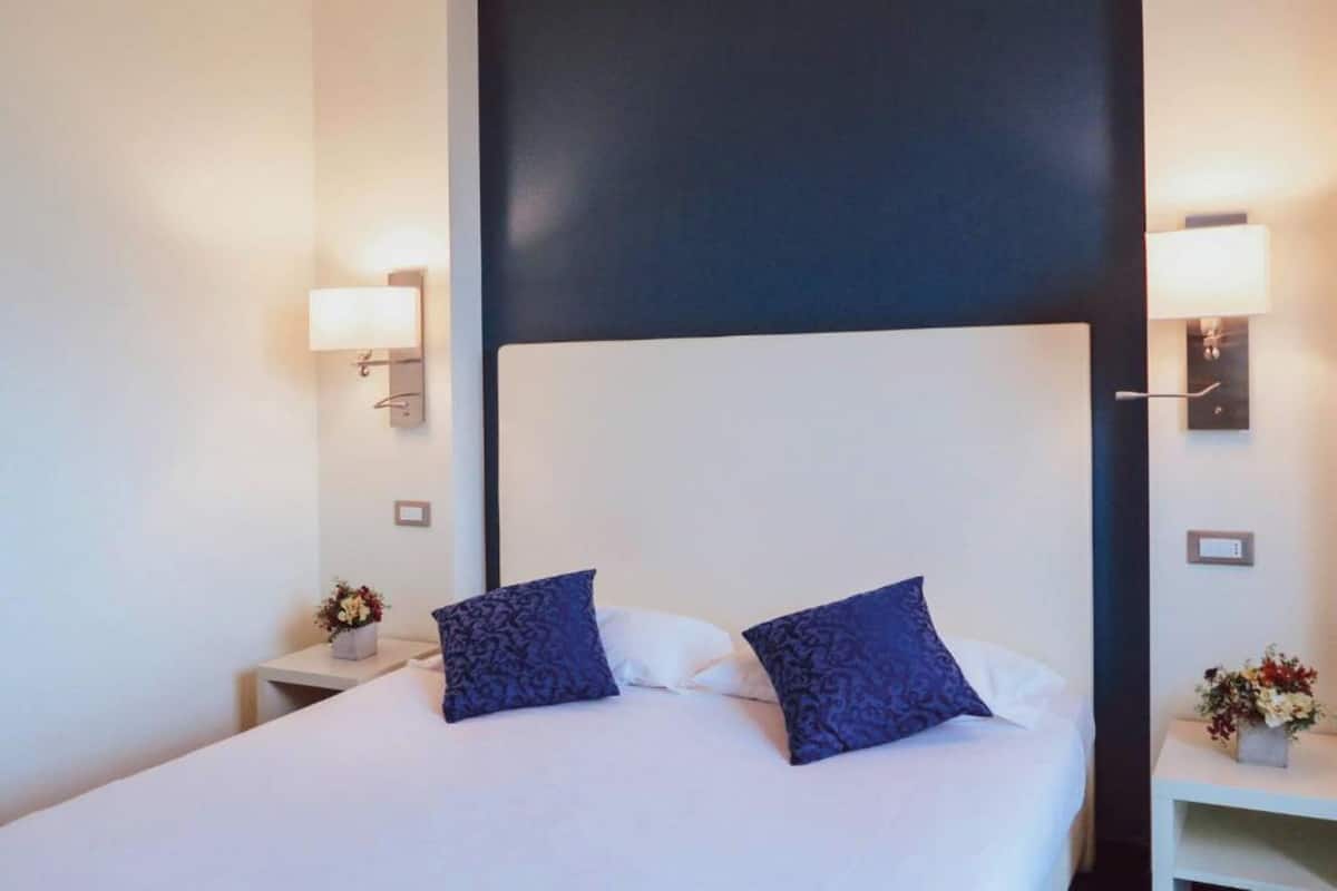 A neatly made bed with white linens and two blue pillows, flanked by bedside tables with lamps and small flower arrangements.