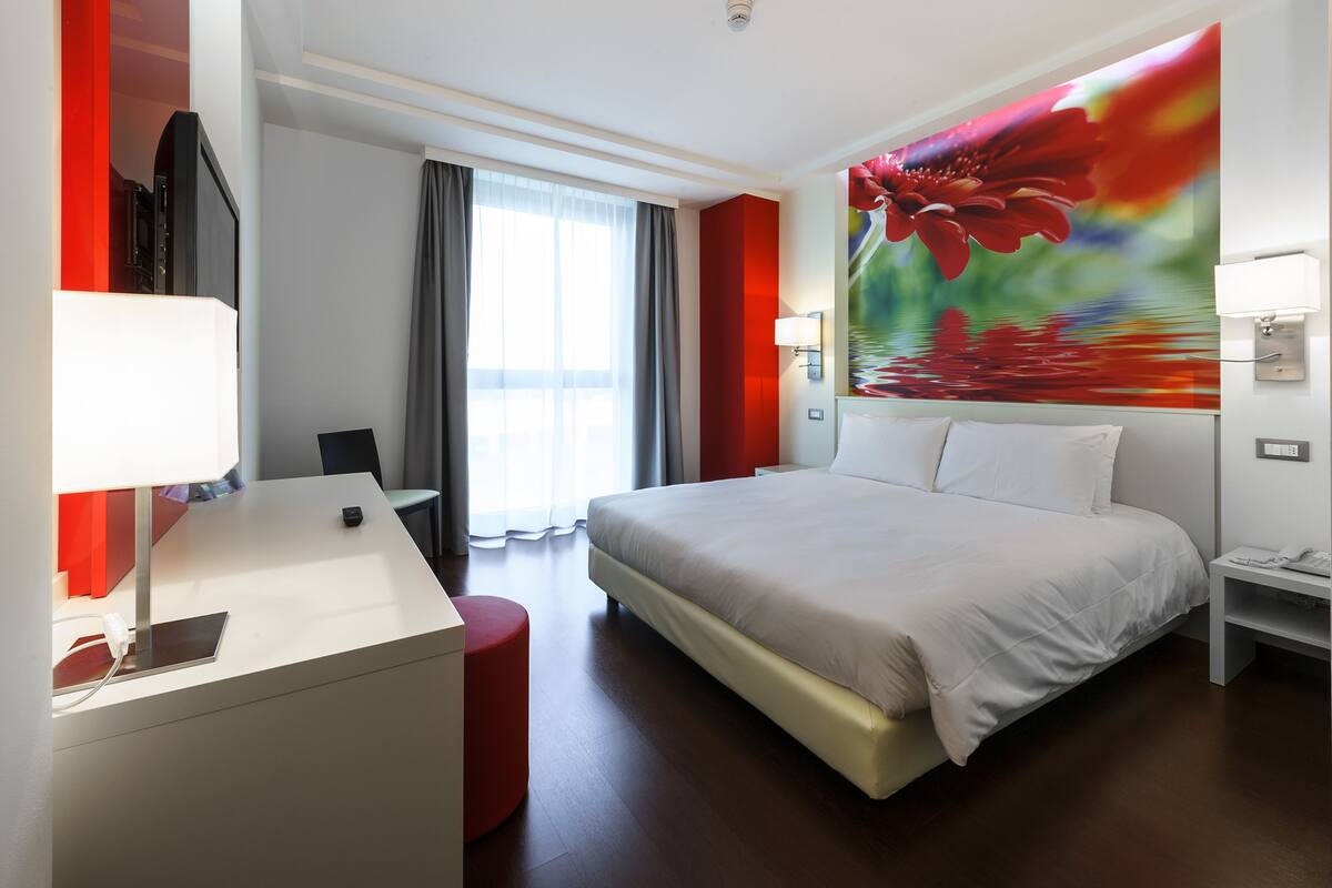 A hotel room with a bed, a desk with a lamp, a TV, a red accent chair, and a large floral mural on the wall.