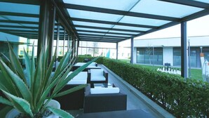 A covered outdoor seating area with white cushioned benches and a large potted plant. - AN HOTEL MILANO MELEGNANO (Carpiano)