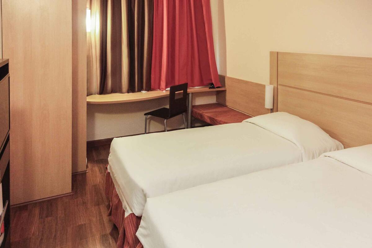Ein Hotelzimmer mit zwei Betten, einem Schreibtisch, einem Stuhl und einem fensterbelaubten Fenster.