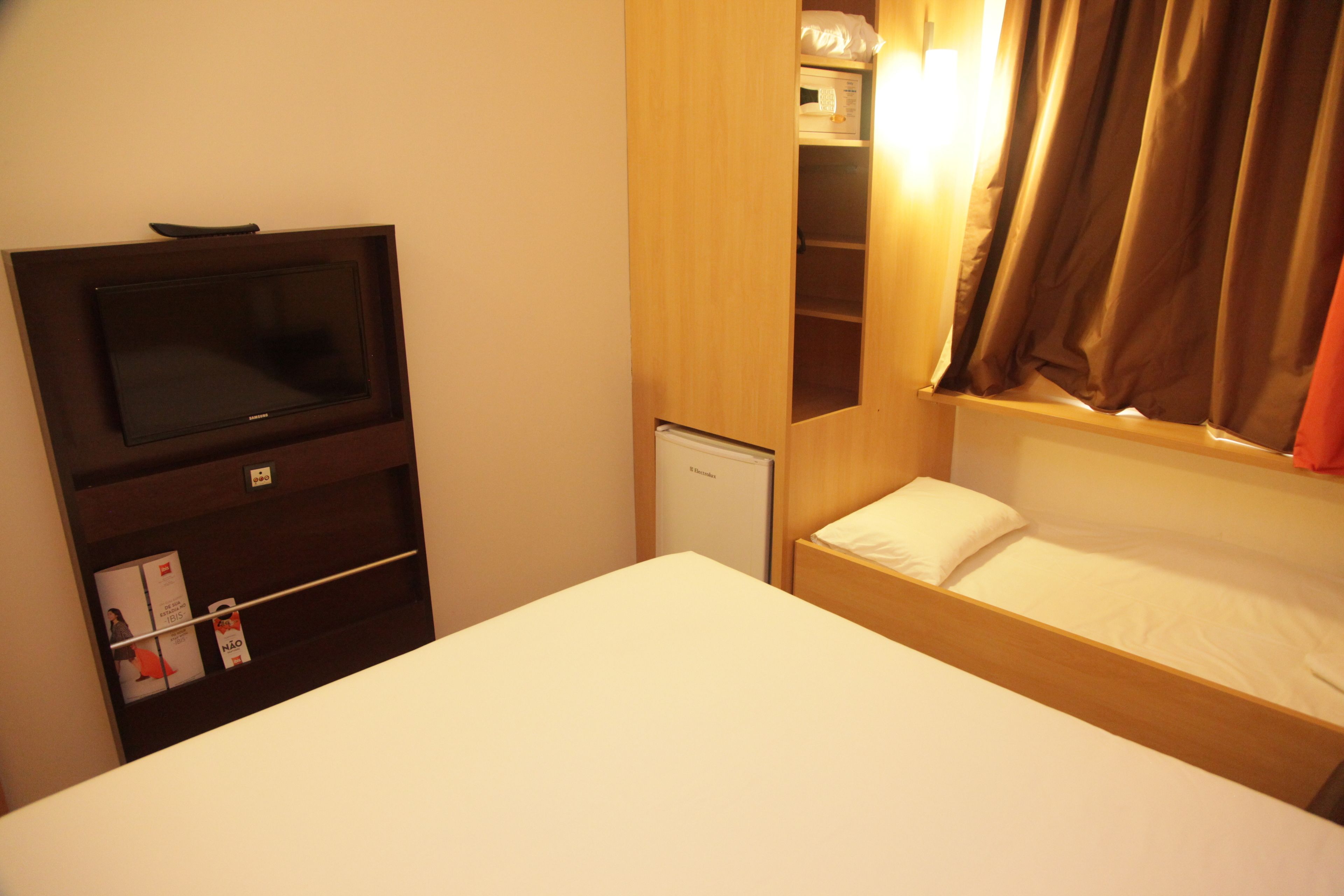 A compact hotel room with a single bed, a small refrigerator, a television mounted on a wooden cabinet, and a window with curtains.
