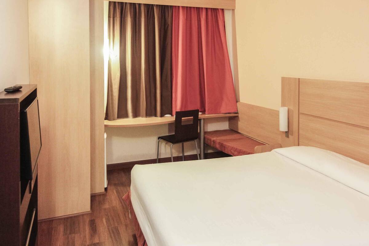Ein Hotelzimmer mit einem Bett, einem Schreibtisch mit Stuhl, einem Fenster mit Vorhängen und einem Fernseher auf einem Tisch.