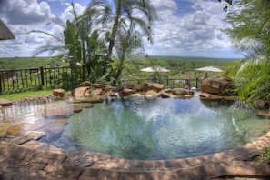 A clear swimming pool with a brick border, surrounded by rocks and palm trees, with a view of the landscape. - Victoria Falls Safari Lodge (Victoria Falls)