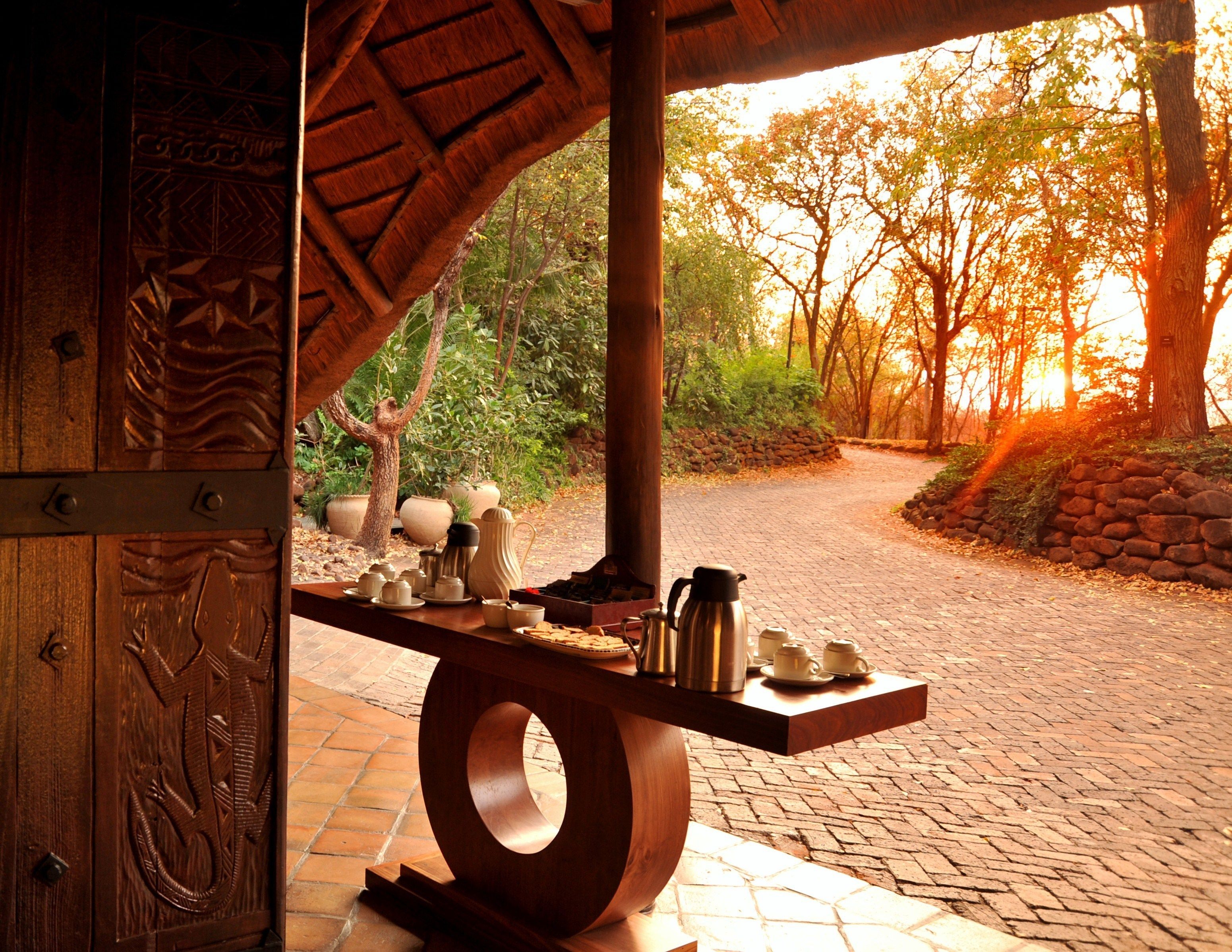 A wooden table with tea sets and a kettle, set against a backdrop of a brick pathway and trees.