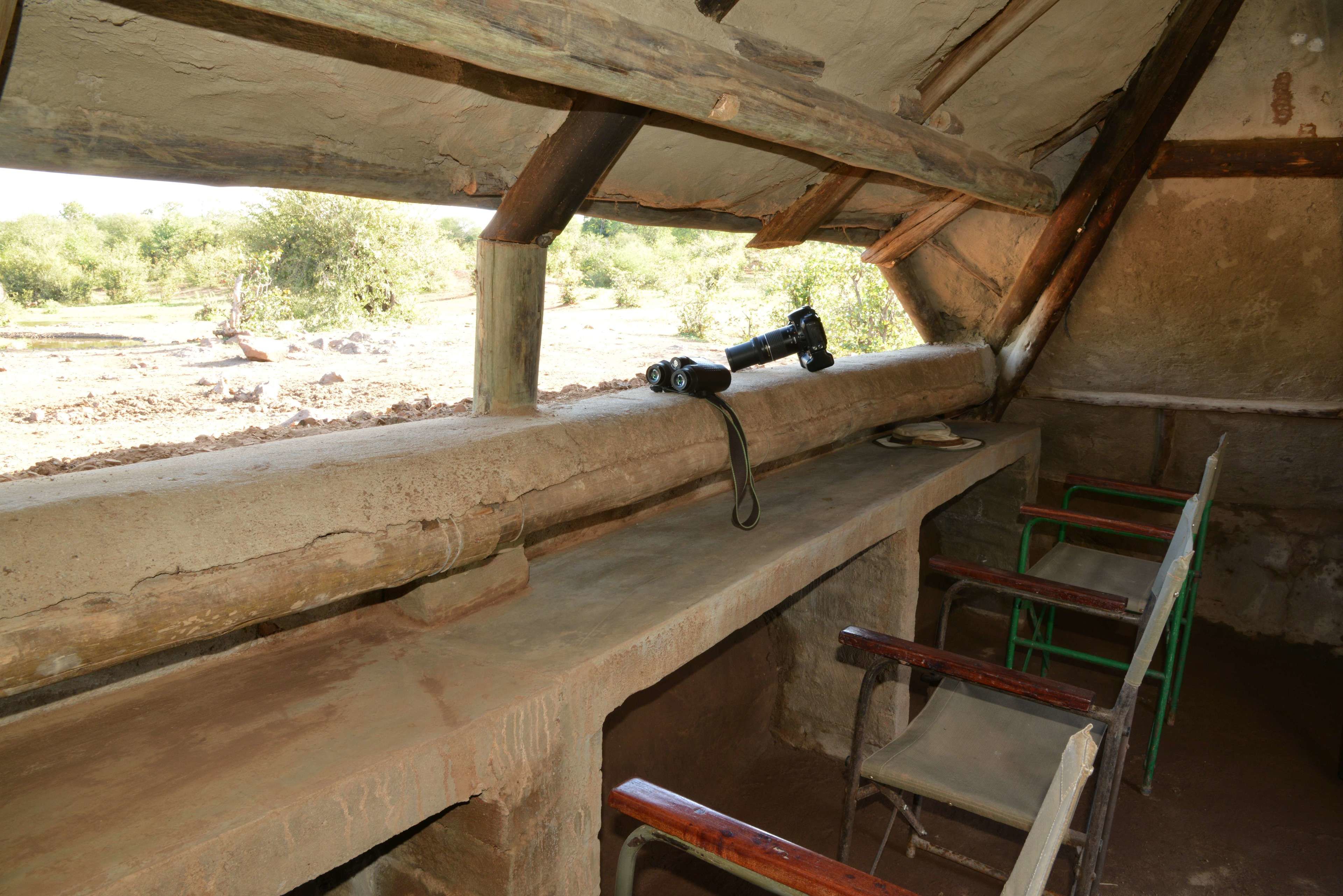 A rustic wooden bench with a camera on it, a green metal chair, and a view of the outdoors.