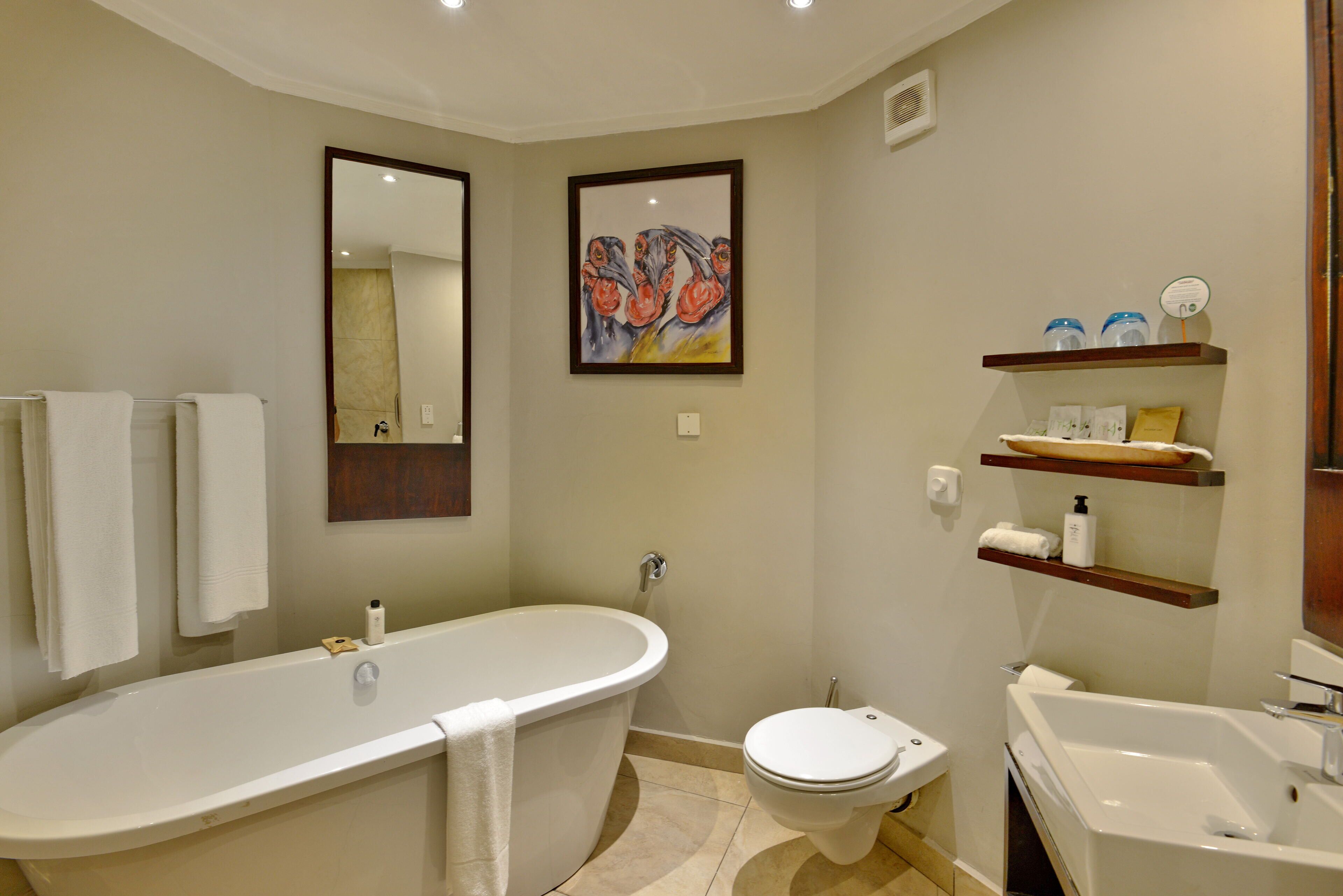 Ein Badezimmer mit freistehender Badewanne, Toilette und Waschbecken. Handtücher hängen an einem Wäscheständer, ein gerahmtes Bild ist an der Wand und es gibt Ablagen mit Pflegeprodukten.