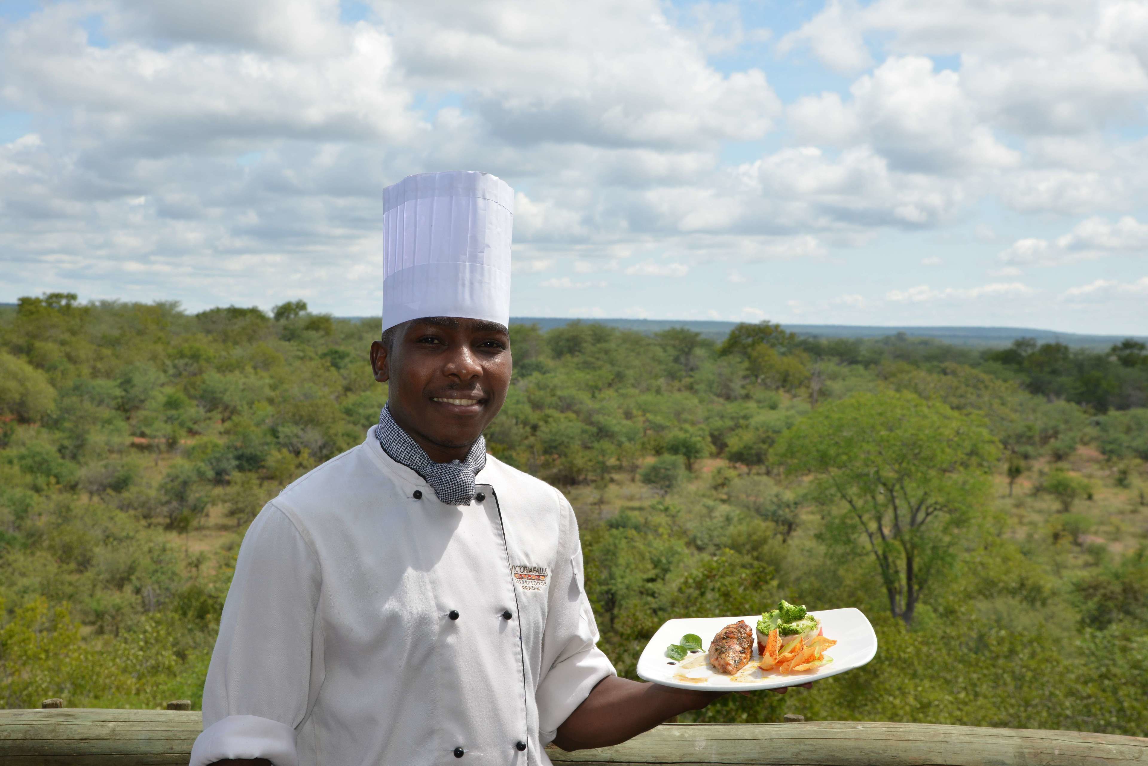 A chef in a white uniform holding a plate of food with a scenic landscape in the background.