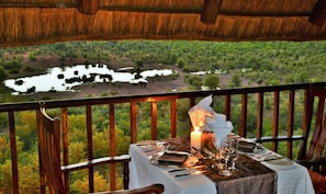 A dining table set for two with a candle, glasses, and cutlery, overlooking a scenic landscape with a water body and trees. - Victoria Falls Safari Lodge (Victoria Falls)
