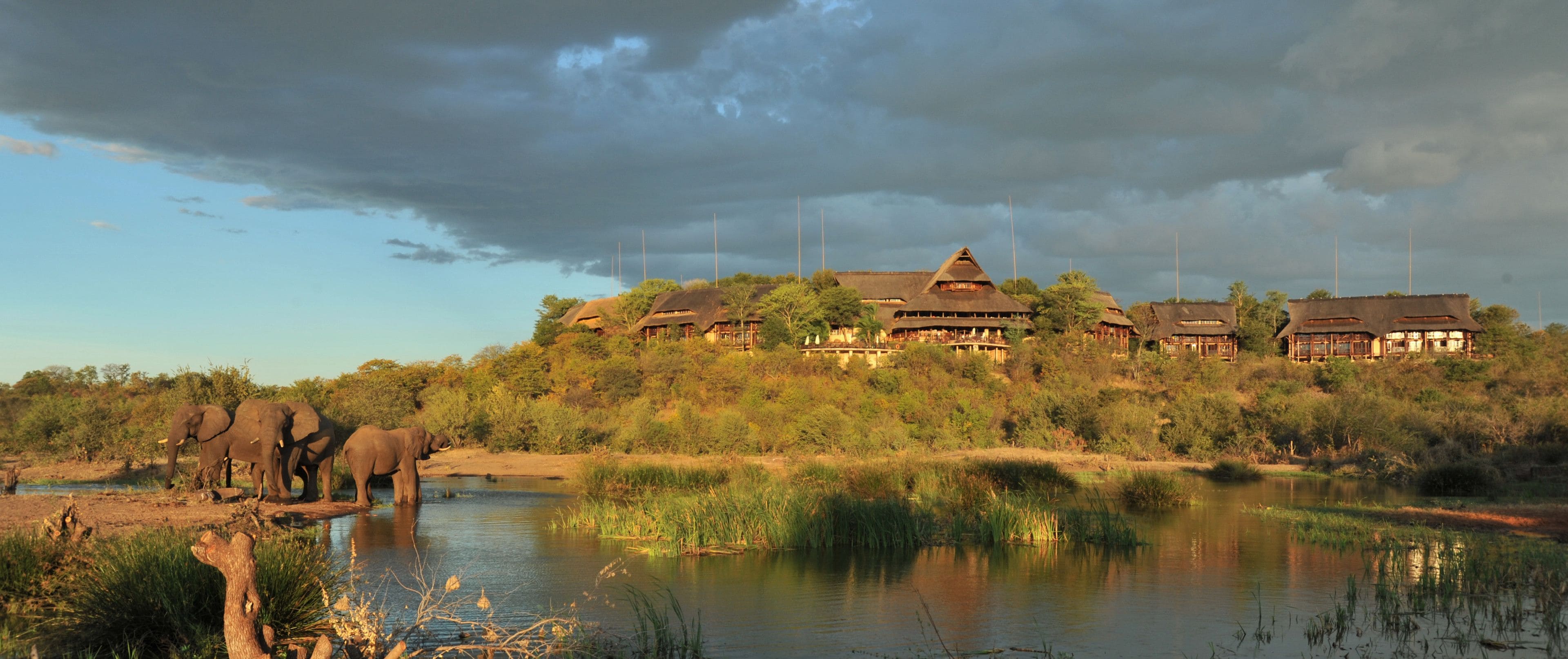 A large building with multiple structures on a hill, surrounded by greenery and a body of water.