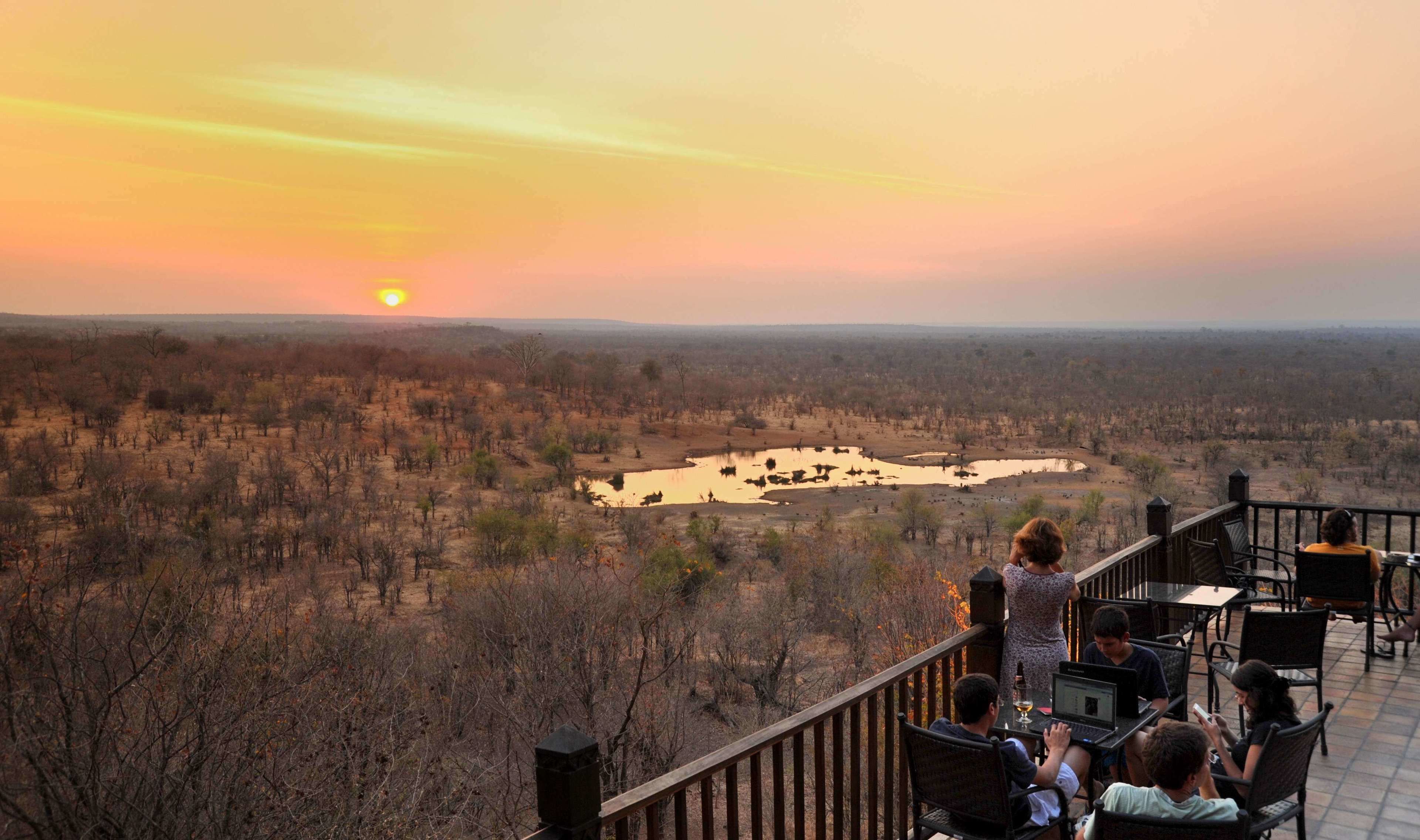 Ein Sonnenuntergangsblick von einer Terrasse über einer trockenen Landschaft mit vereinzelter Vegetation und einem kleinen Gewässer.