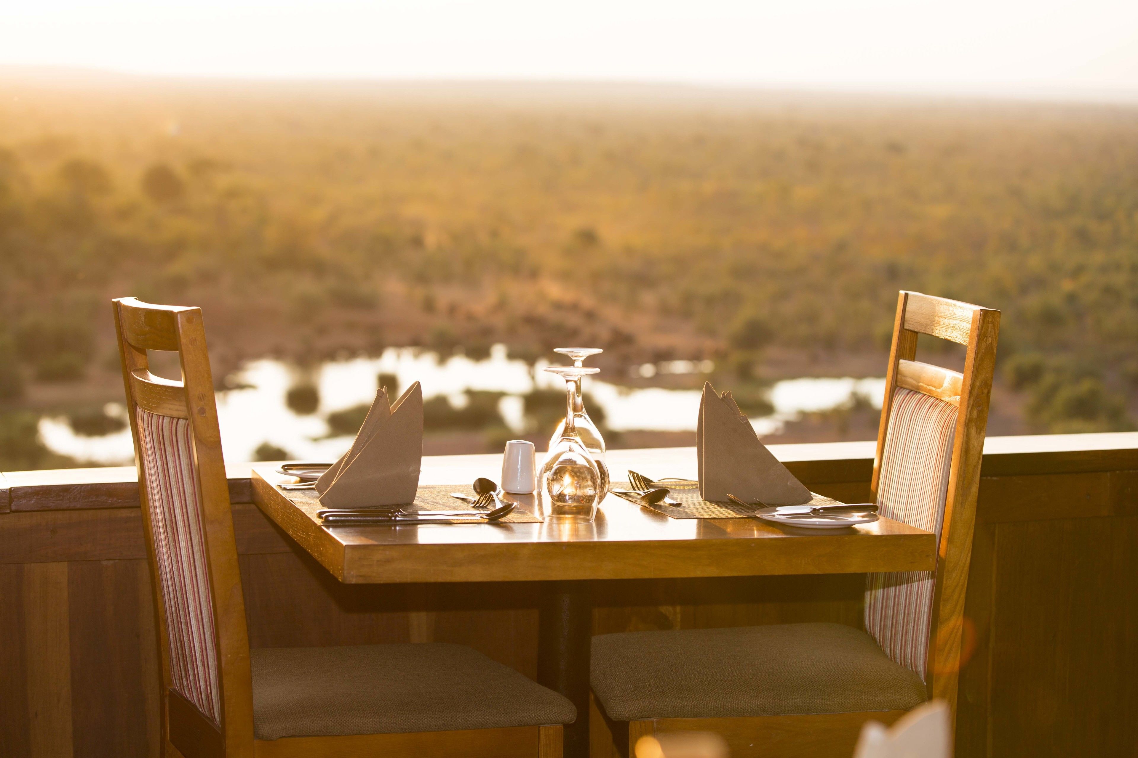 A wooden dining table with chairs, set for two, overlooking a scenic landscape with a body of water and trees.