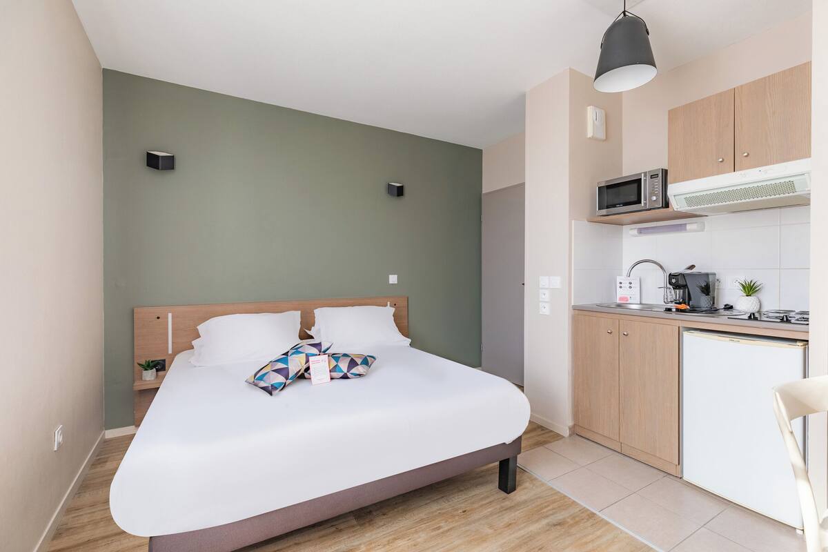 A modern room with a bed, a small bedside table, a microwave, a coffee maker, and a refrigerator.