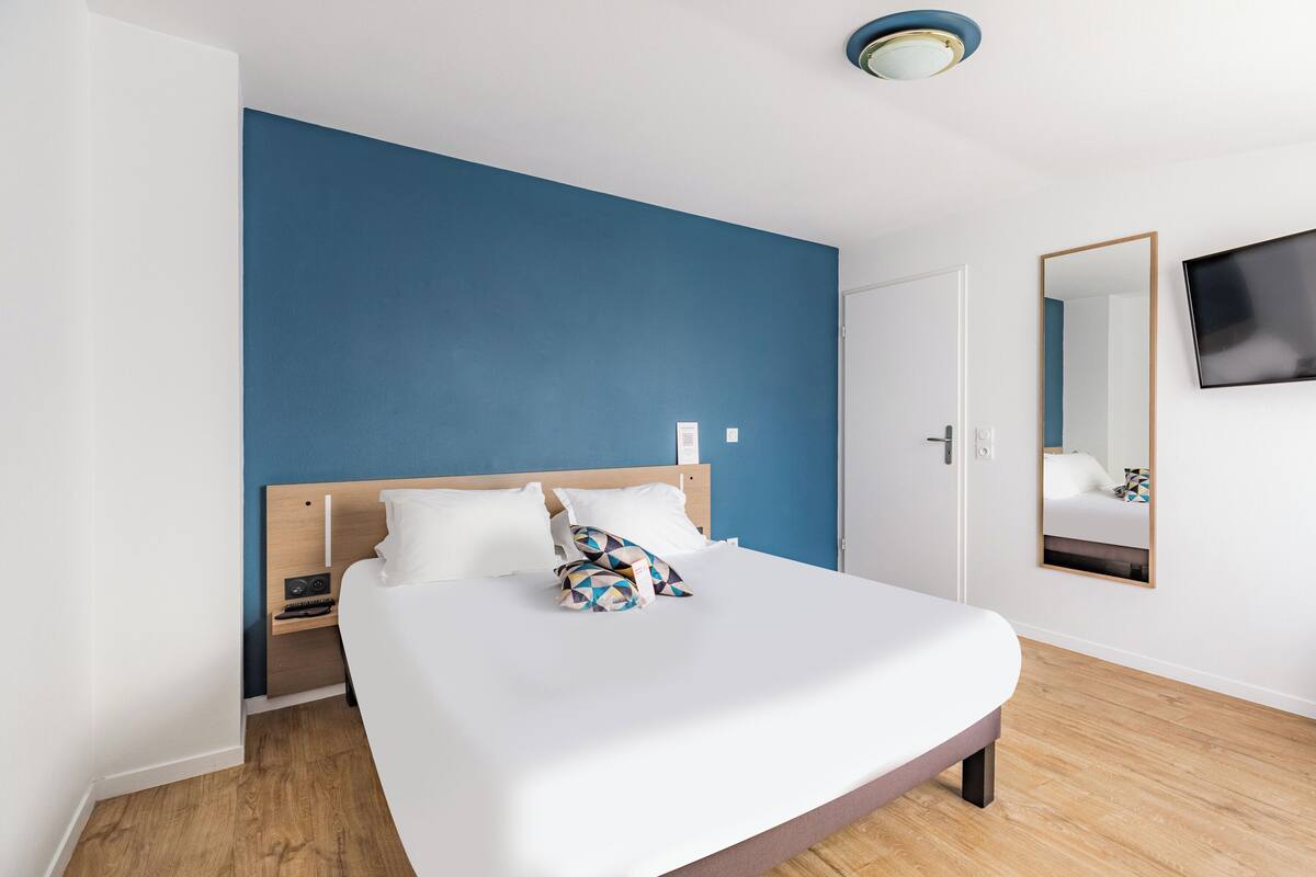 A modern hotel room with a large bed, a wooden headboard, a flat-screen TV mounted on the wall, and a small bedside table.