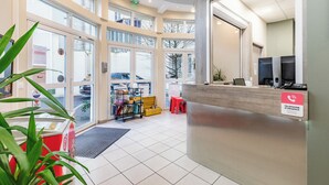 A reception area with a reception desk, a telephone emergency sign, a plant, and a cart with books and a yellow box. - Appart'City Confort Niort Centre (Niort)