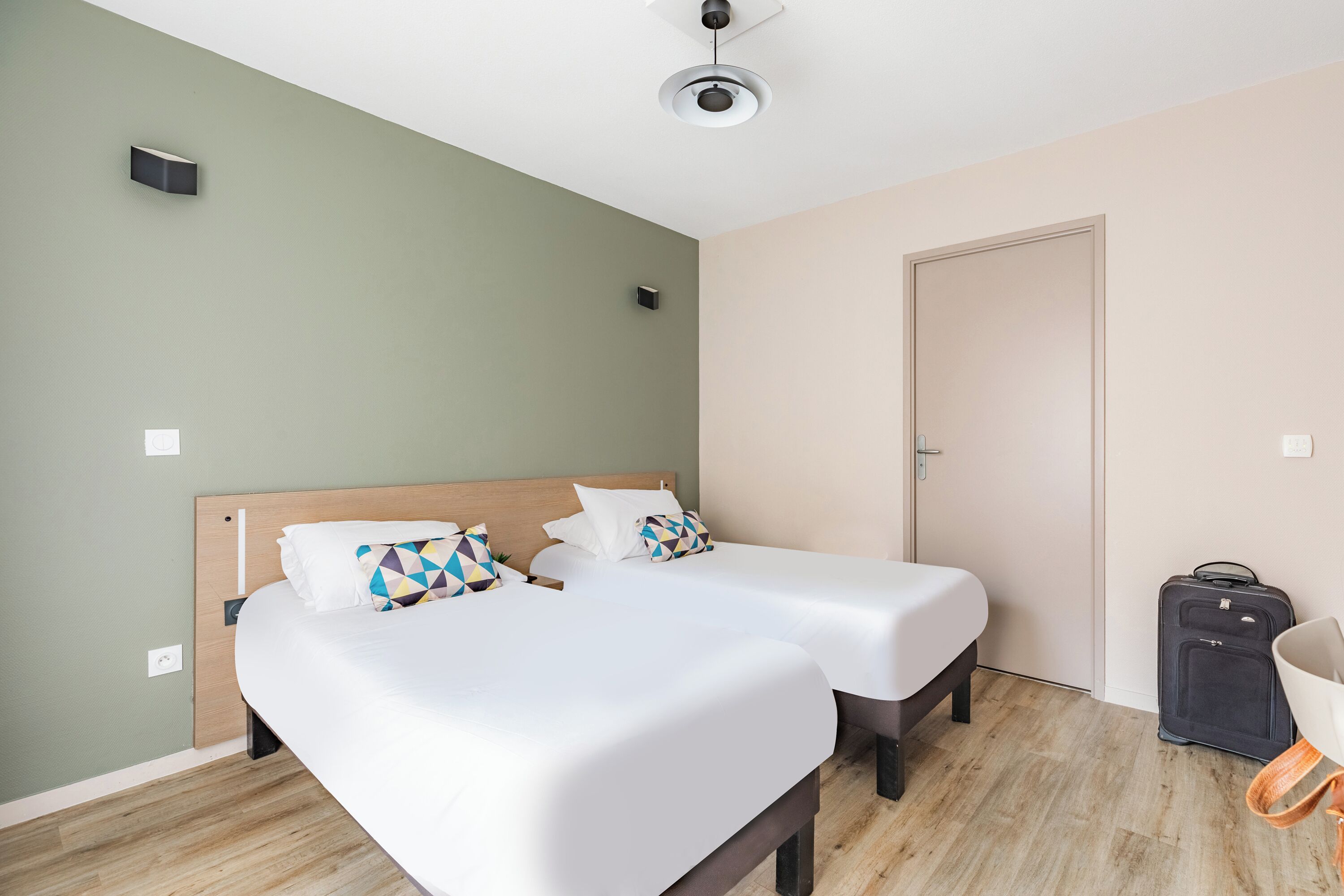 Habitación de hotel con dos camas individuales, un cabecero de madera, un ventilador de techo y una maleta en el suelo.