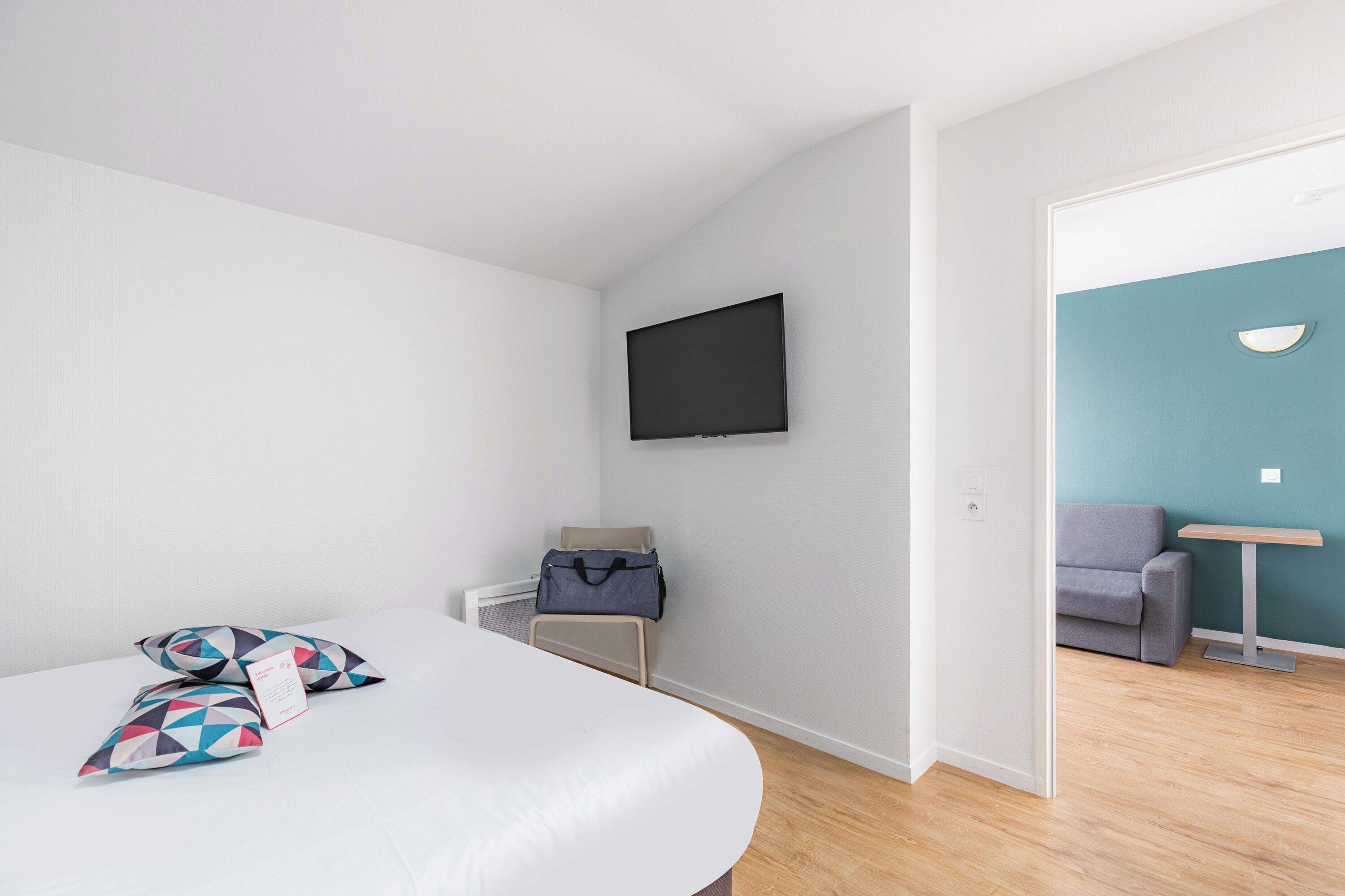 Une chambre d’hôtel moderne équipée d’un lit, d’un téléviseur fixé au mur, d’une petite table et d’un canapé dans la pièce adjacente.