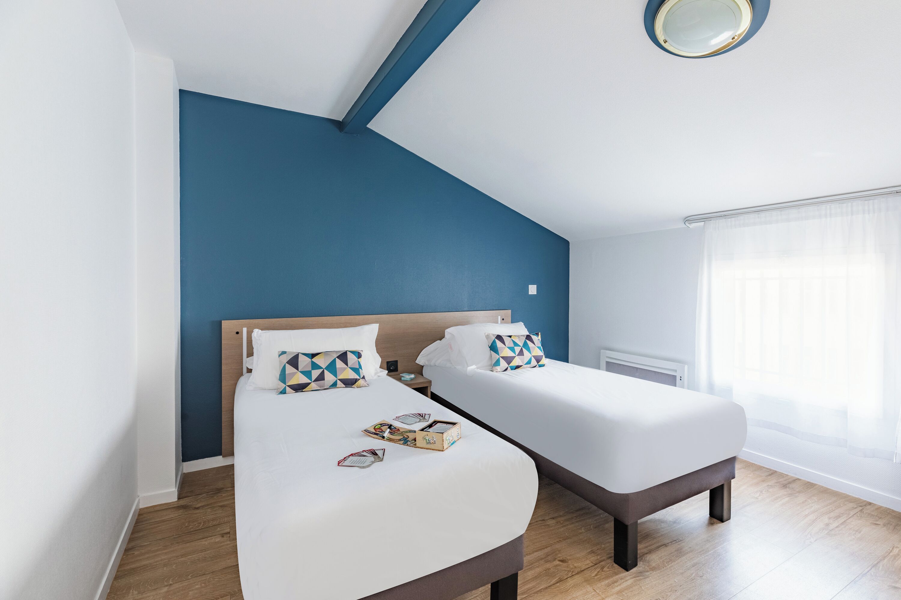 Ett modernt hotellrum med två separata sängar, ett trägolv, en blå accentvägg och ett stort fönster med tunna, genomskinliga gardiner.