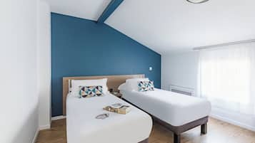 Ett modernt hotellrum med två separata sängar, ett trägolv, en blå accentvägg och ett stort fönster med tunna, genomskinliga gardiner.