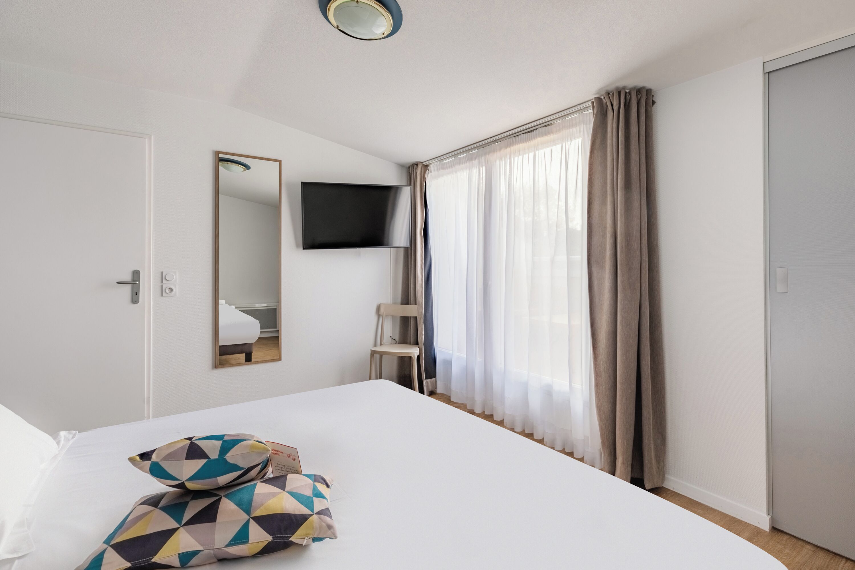 Hotellihuone, jossa on sänky, seinälle asennettu televisio, tuoli ja ikkuna, jossa on verhot.