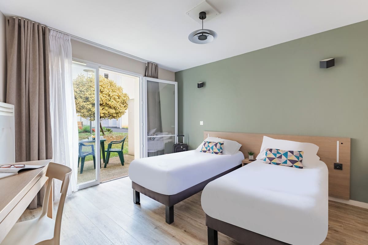 A hotel room with two beds, a desk, and a sliding glass door leading to an outdoor area.