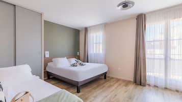 A modern hotel room with a large bed, a smaller bed, a wooden floor, and a window with sheer curtains.