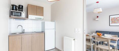 Una cocina moderna con armarios de madera, un refrigerador blanco y un microondas. Hay un comedor con una mesa y sillas, y un radiador en la pared.