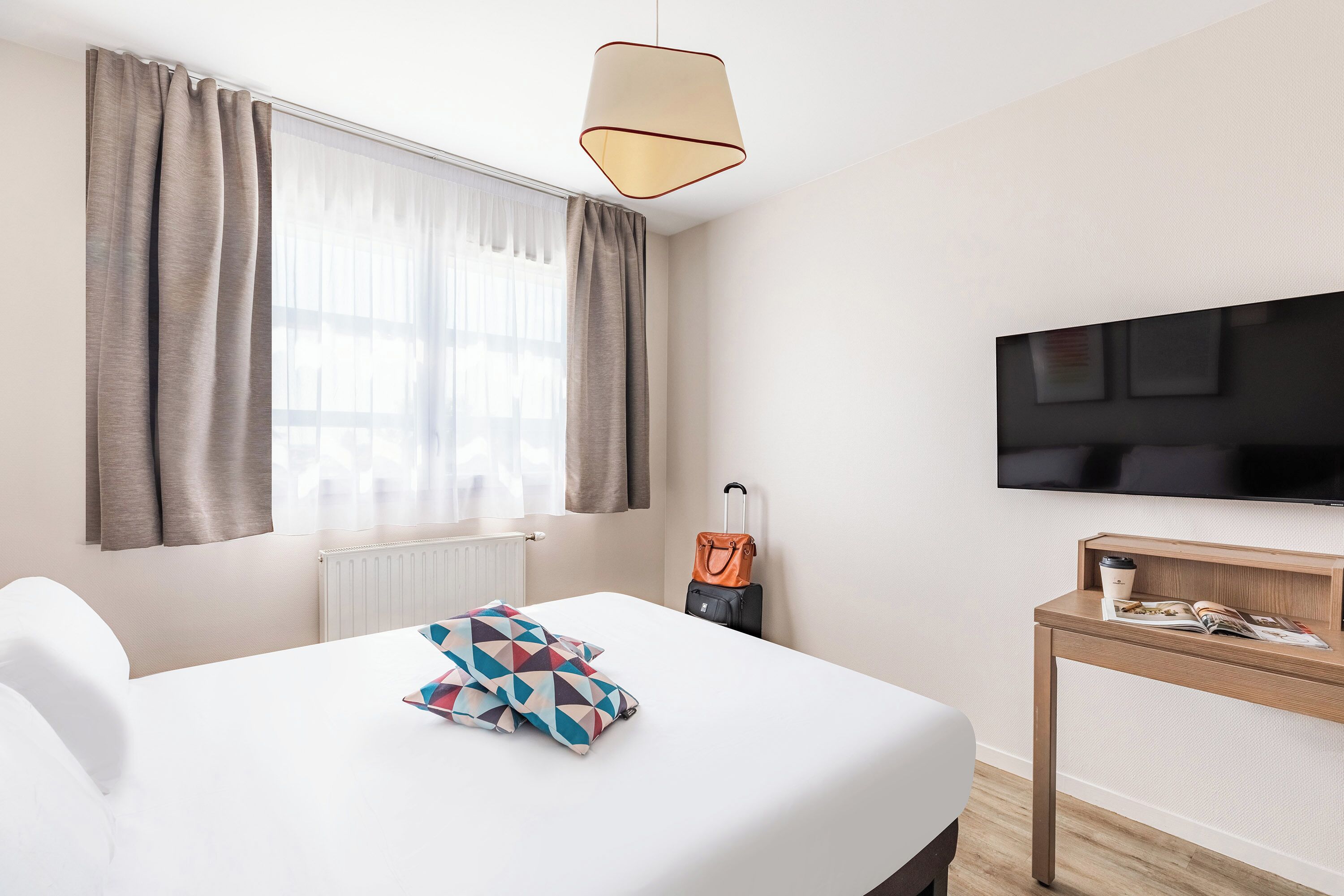 Une chambre d’hôtel avec un lit, un téléviseur fixé au mur, une table de chevet en bois et une fenêtre avec des rideaux.