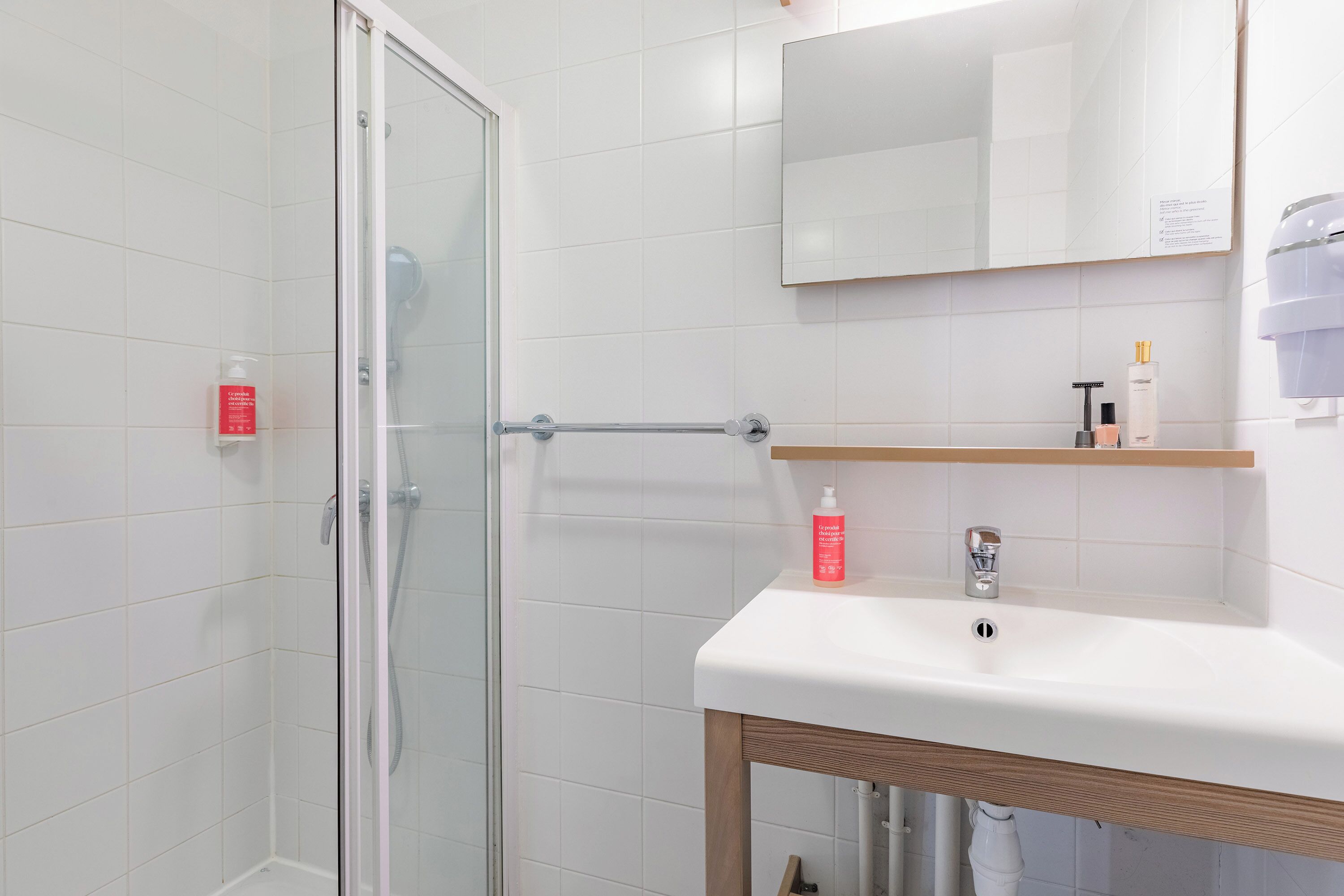A modern bathroom with white tiles, a glass shower enclosure, a wooden-framed sink, and a mirror above it.