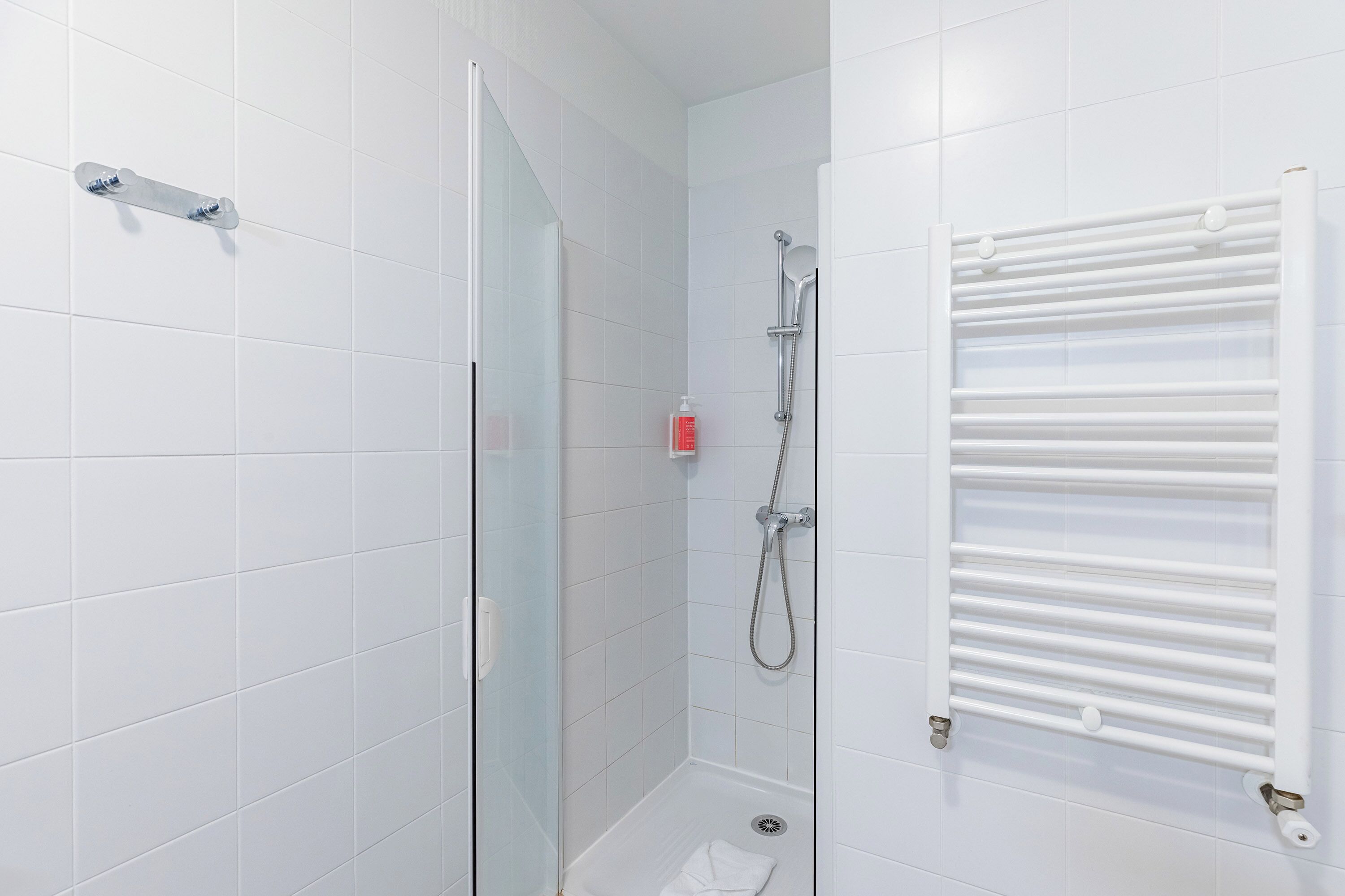 A bathroom with white tiled walls, a glass shower door, a white towel on the floor, and a white towel rack.