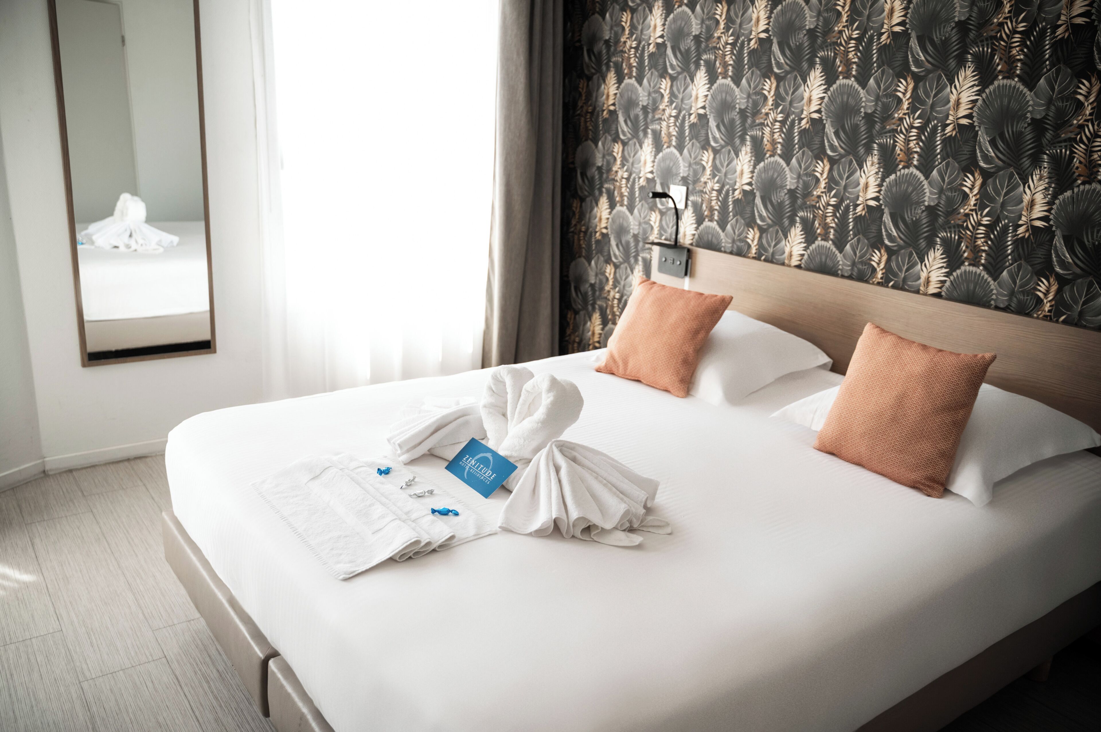 A hotel room with a neatly made bed, a mirror, and a wall with a leaf pattern.
