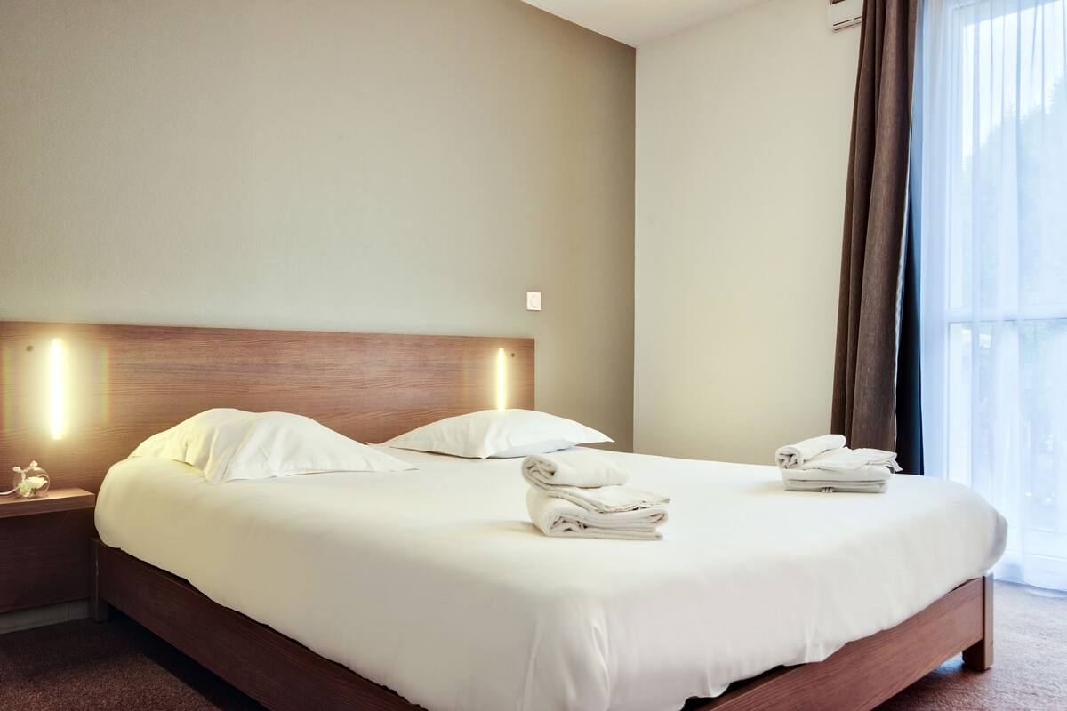 A neatly made bed with white linens and pillows, a wooden headboard, and a bedside table with a lamp and a small decorative item.
