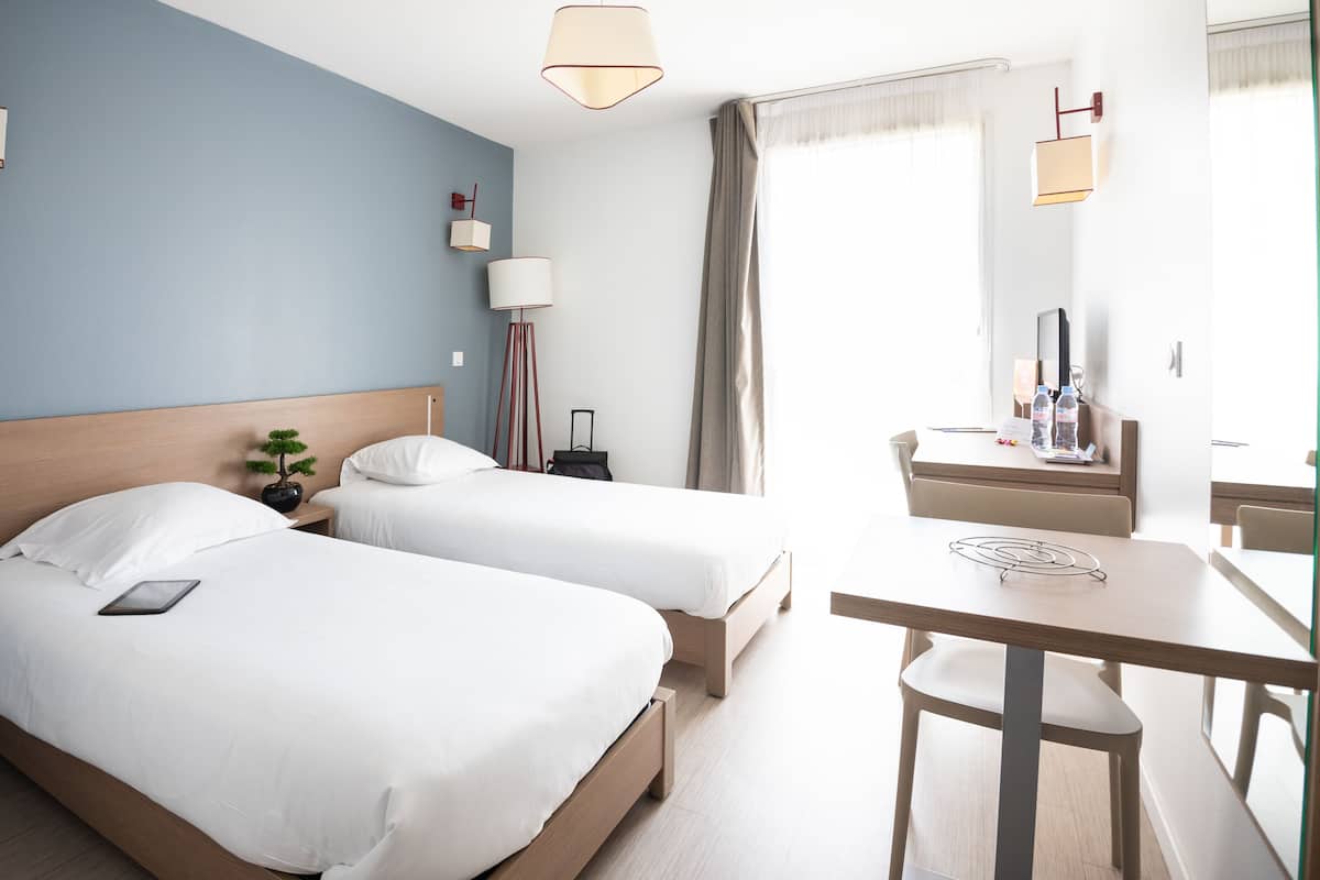 A hotel room with two beds, a desk, a chair, a lamp, and a small plant.