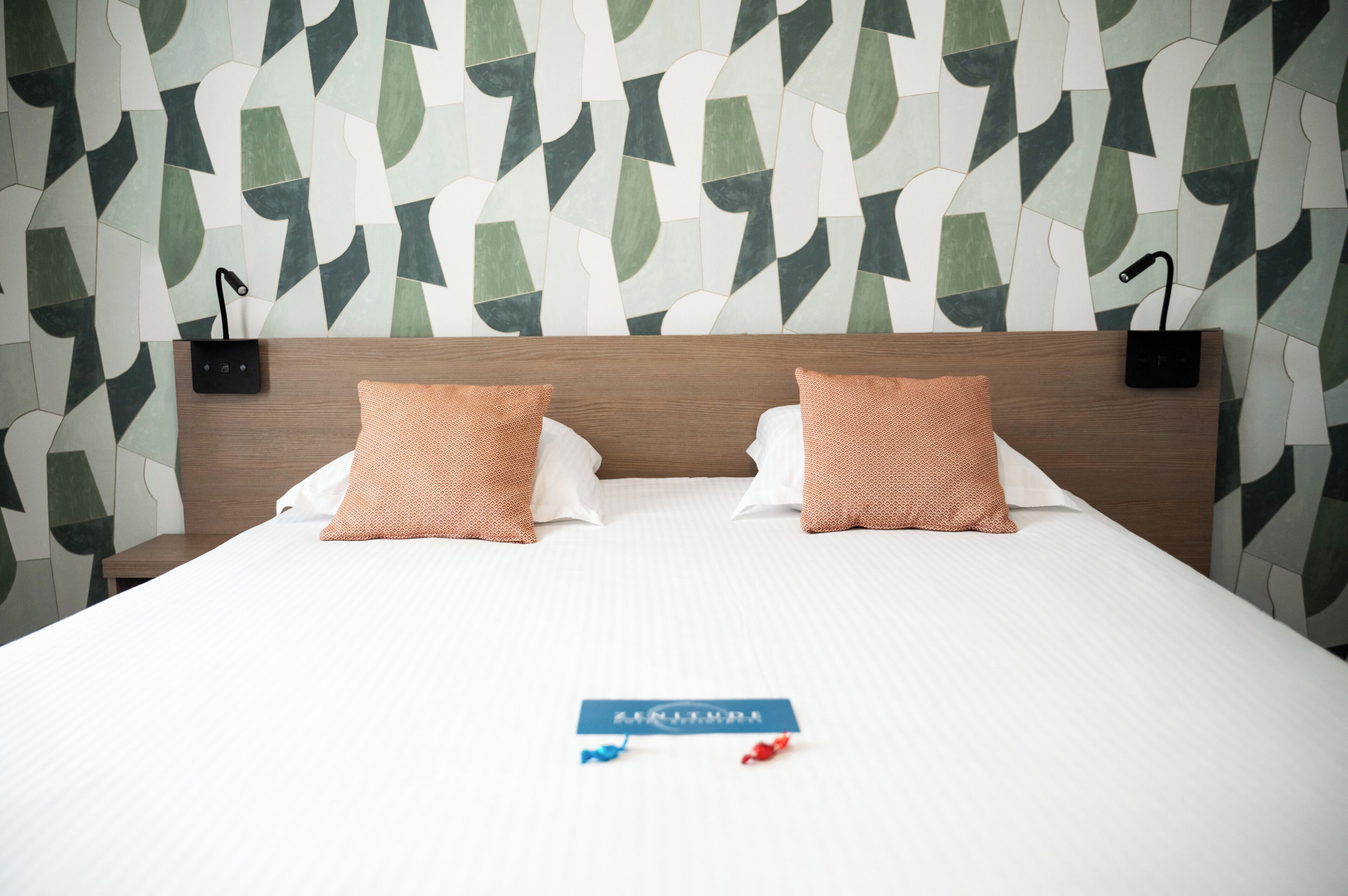 Habitación de hotel con cama, dos almohadas, una mesita de noche y una pared con diseño geométrico.