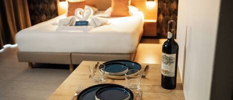 Un lit bien fait, avec des oreillers, une table de chevet sur laquelle on trouve des assiettes, des couverts et une bouteille de vin.