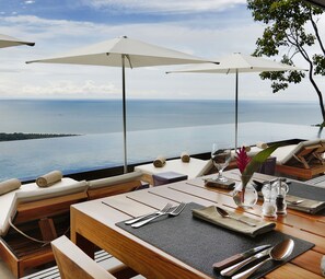 A dining area with a view of the ocean, equipped with wooden tables, white umbrellas, and a glass vase with flowers.