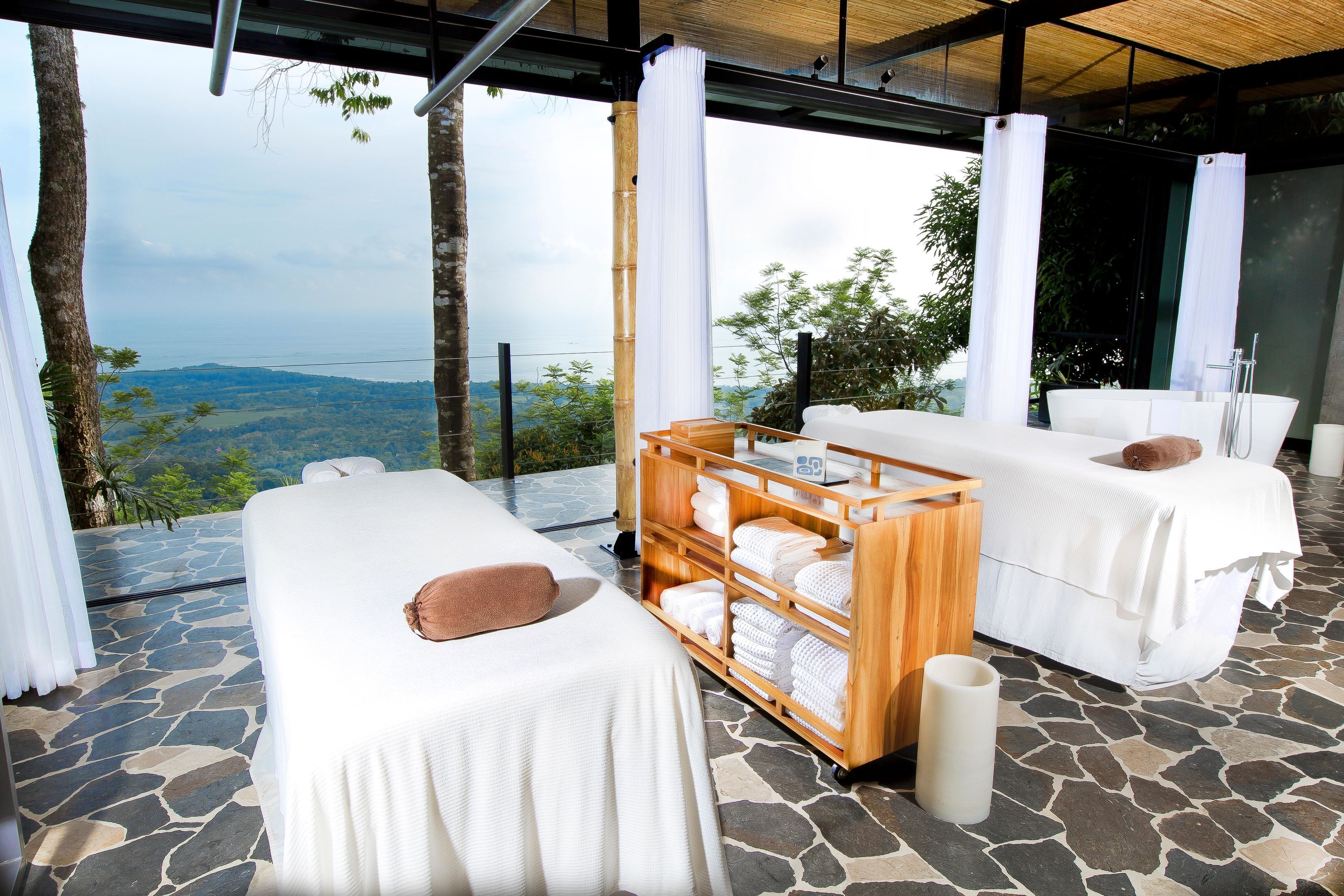 A spa room with two massage tables, a wooden shelf with towels, and a view of the landscape.