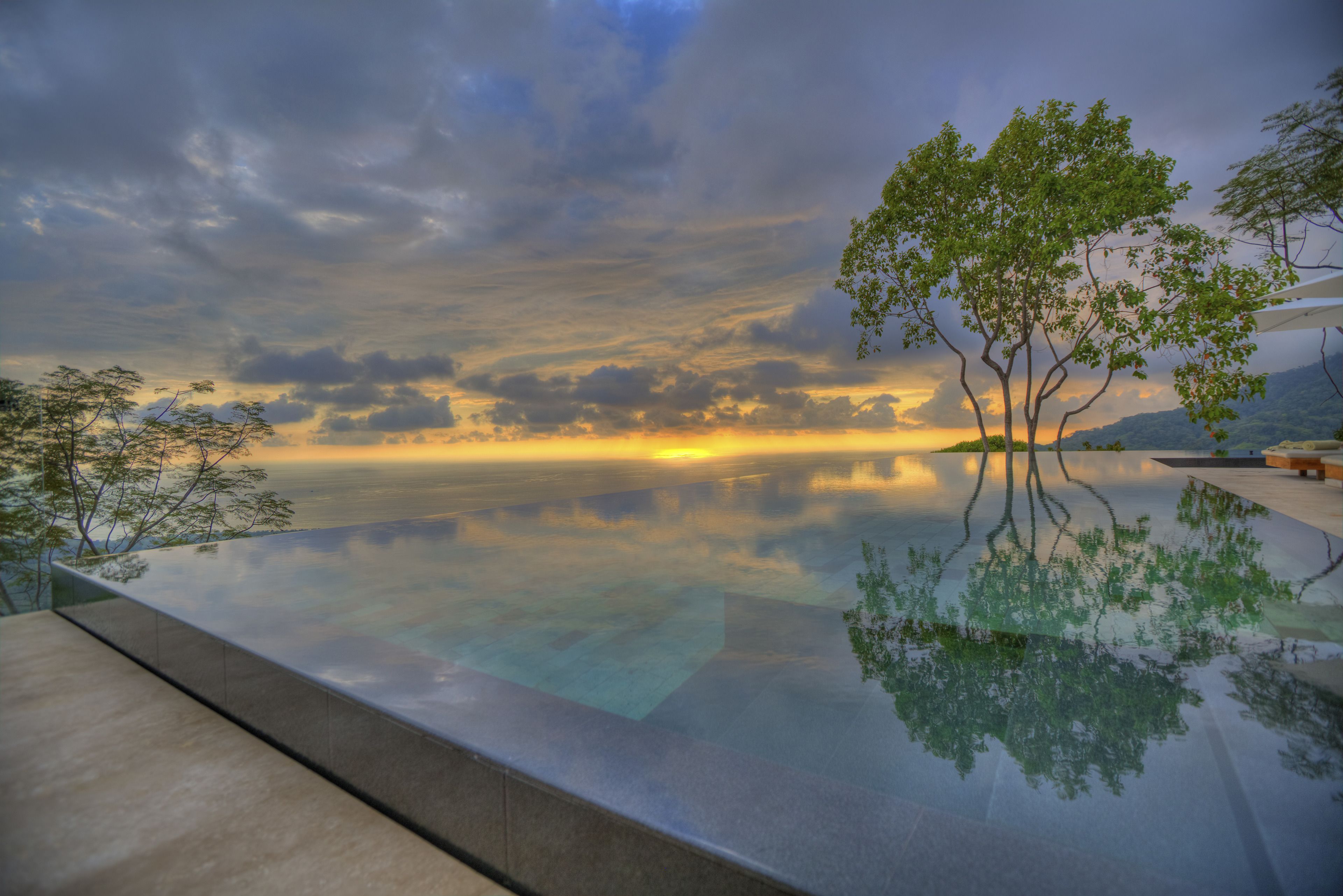 A serene infinity pool reflecting a sunset with a tree in the foreground.