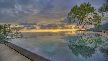 A serene infinity pool reflecting a sunset with a tree in the foreground.